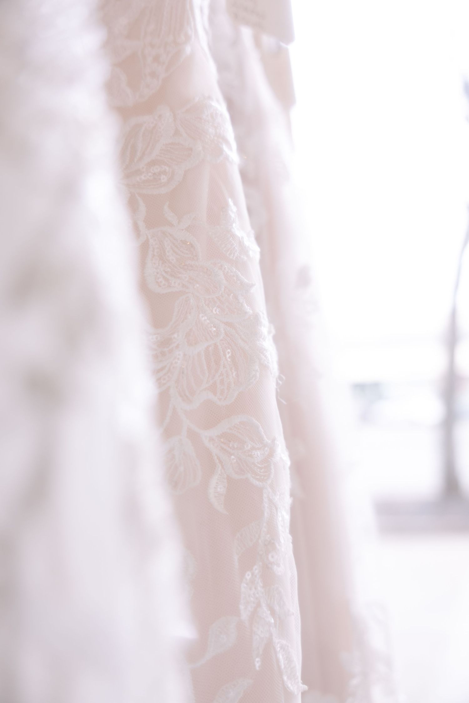 Close-up of elegant white lace wedding dress with intricate floral embroidery details hanging in soft natural light.