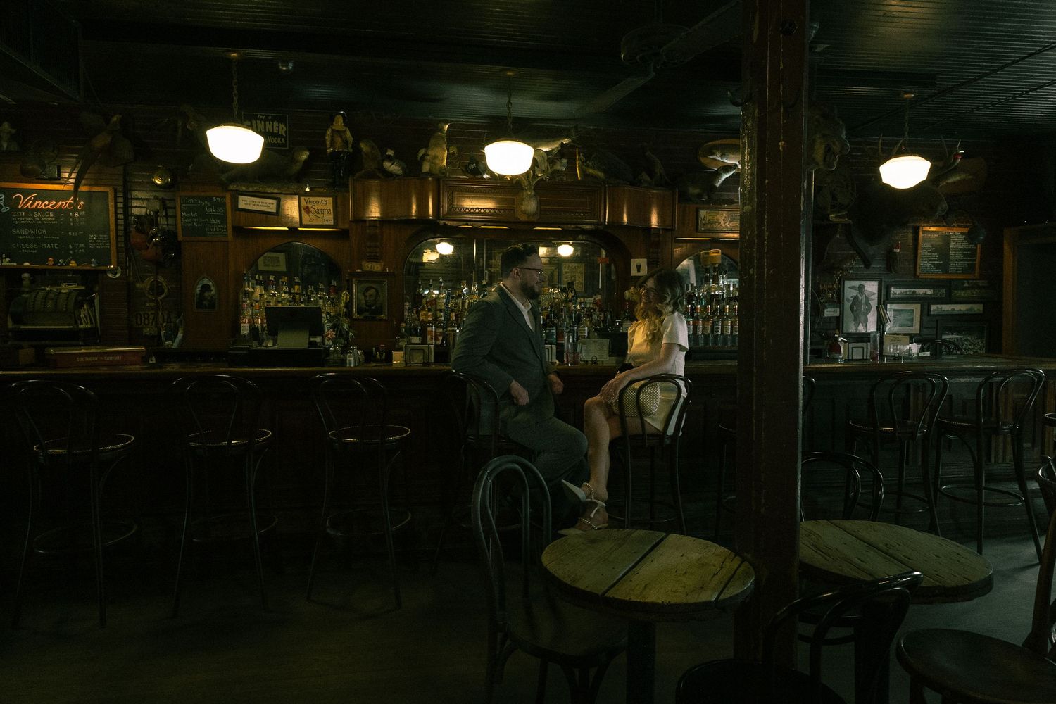 Dimly lit vintage bar interior with two patrons seated at the counter amid dark wood decor and hanging lights.