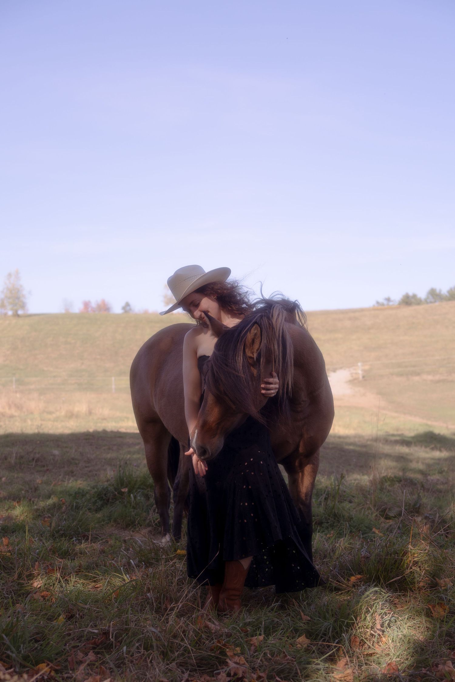 A person in black clothing stands next to a large brown horse on a grassy hillside at sunset.