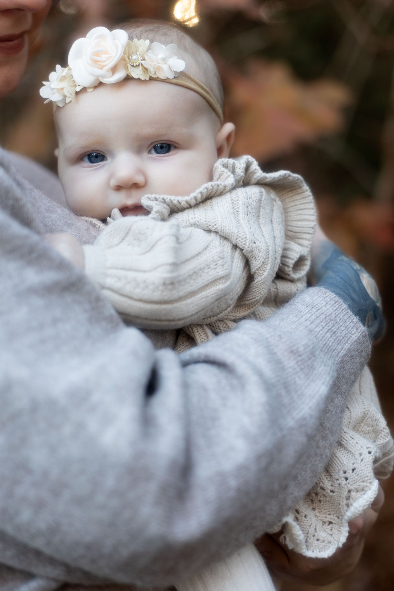 A baby wearing a white lace dress and headband looks to the side while bundled in a cozy gray sweater.