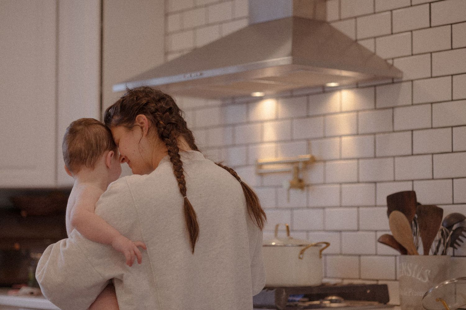 A parent and child share a tender moment in a modern white-tiled kitchen with stainless steel appliances.
