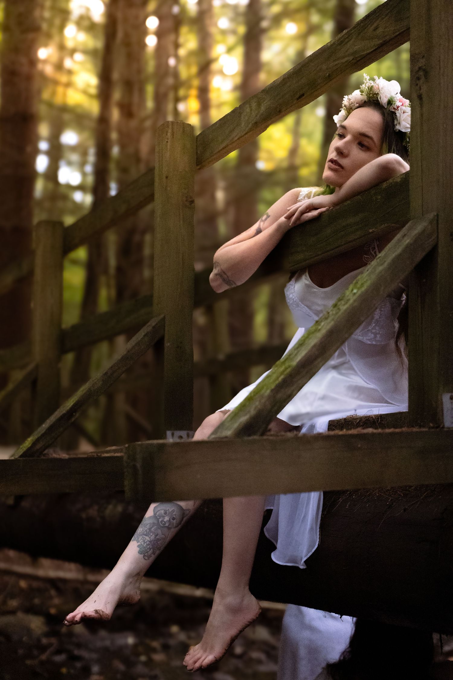 A person in white clothes reclines against a rustic wooden fence in a sunlit forest setting.