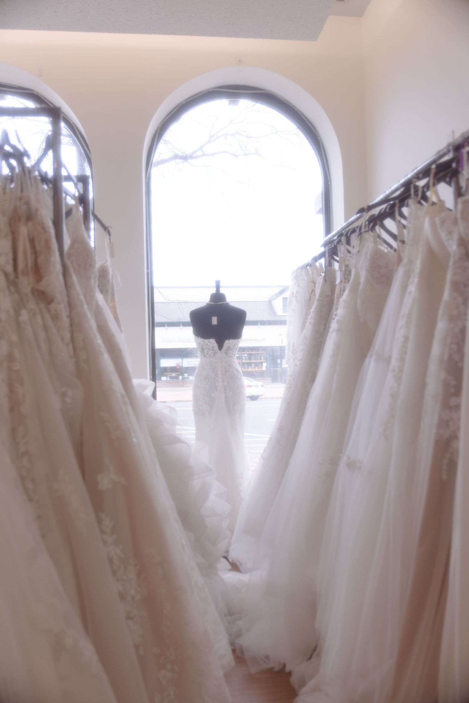 Bridal boutique interior featuring rows of white wedding gowns on racks with a dress displayed on mannequin by arched window.