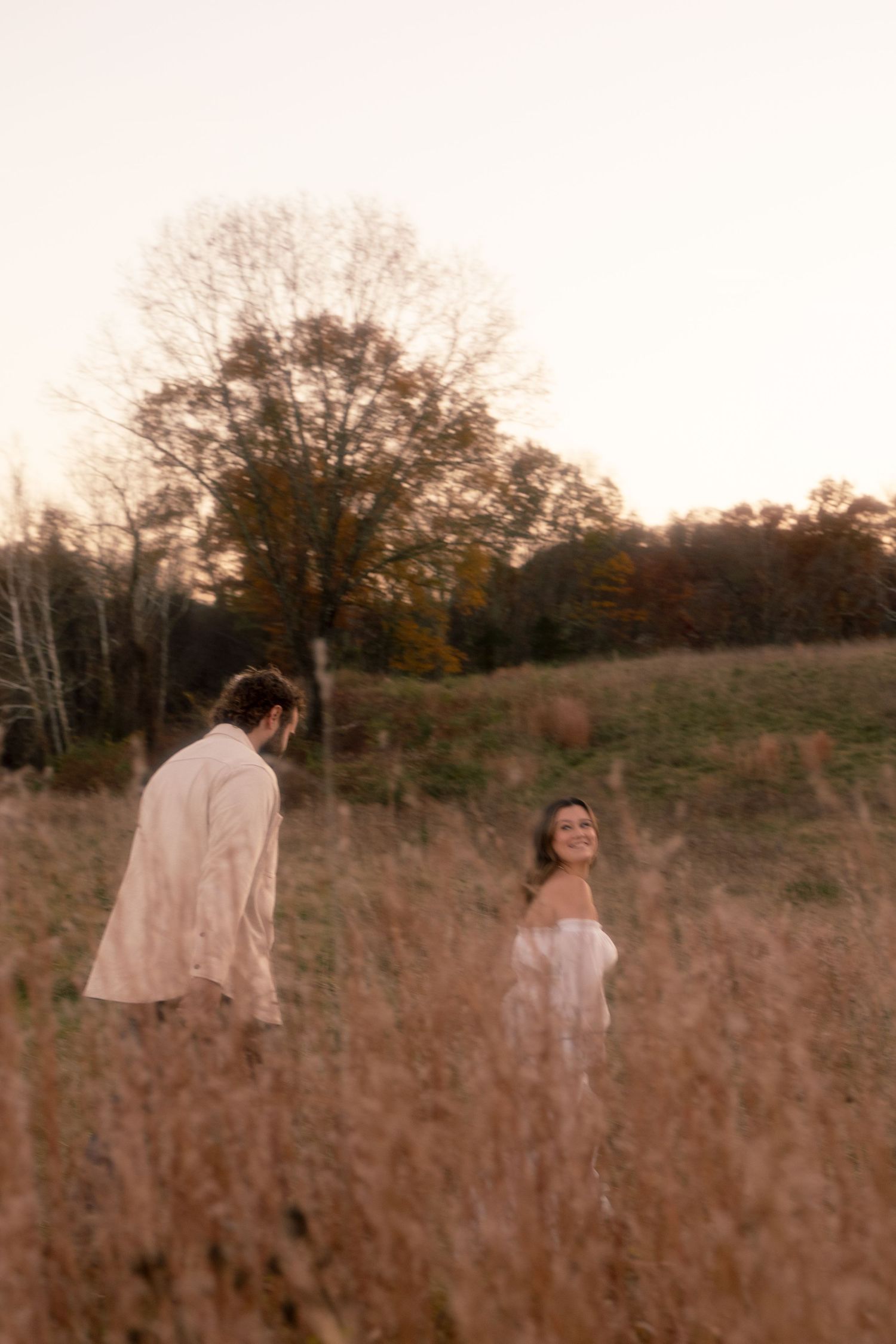Two figures walk through a sepia-toned field of tall grass at sunset with trees in the background.