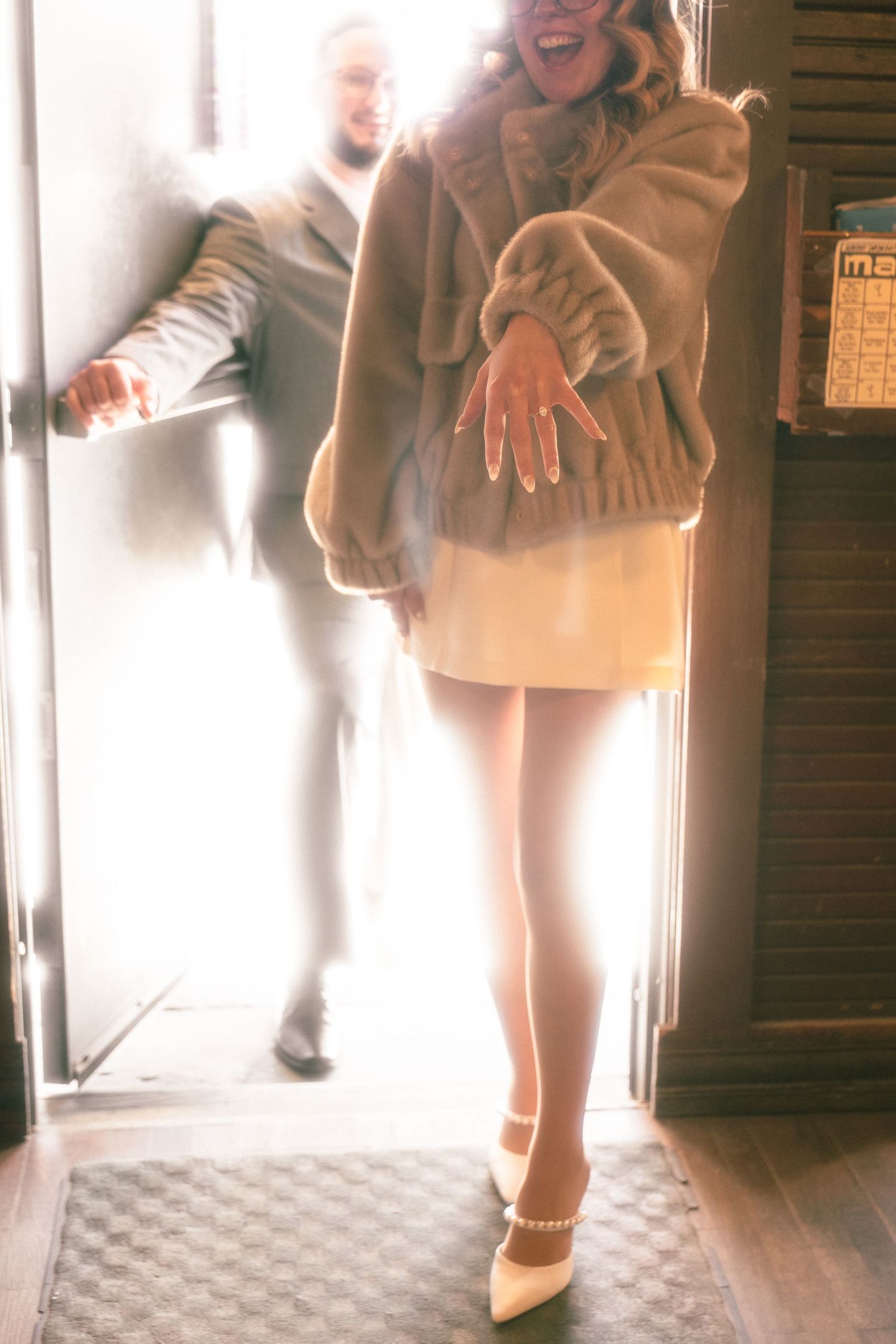 Woman in fur coat and white mini skirt showing engagement ring while entering a bright doorway with man behind her.