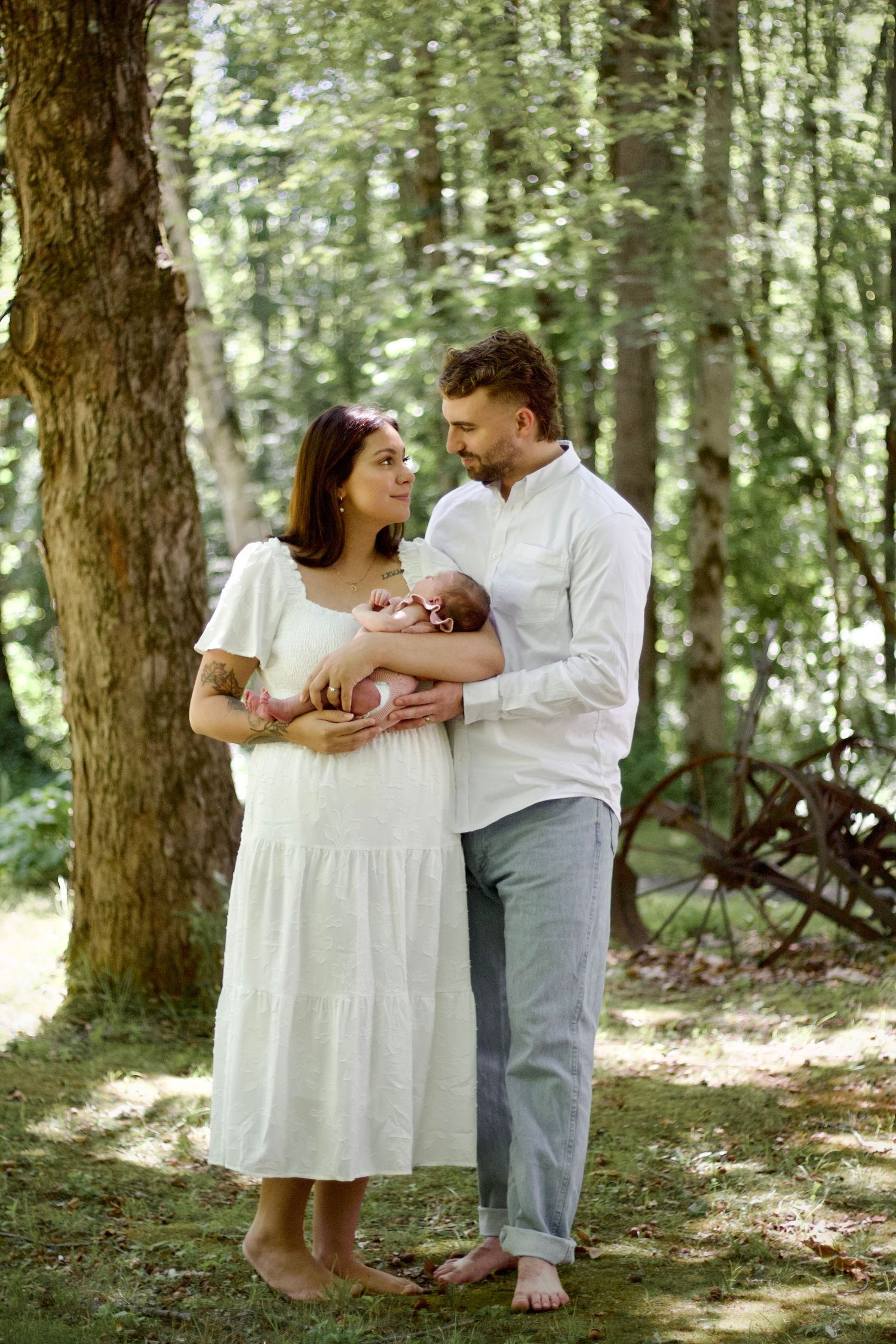 A couple stands barefoot in a peaceful forest setting holding their infant child in summer sunlight.