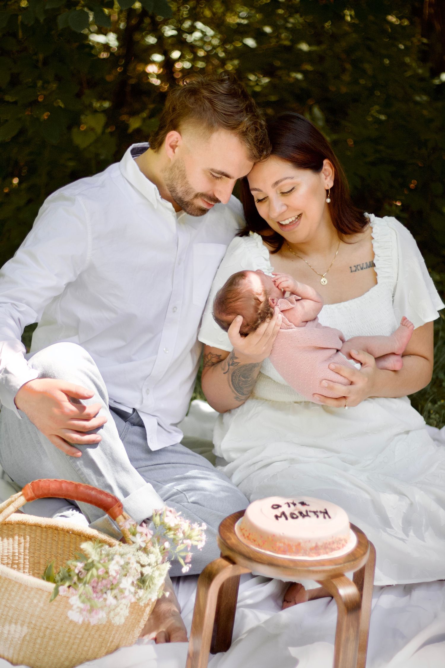 A couple sits on a blanket at an outdoor picnic with a small baby and pink cake on a wooden stool.