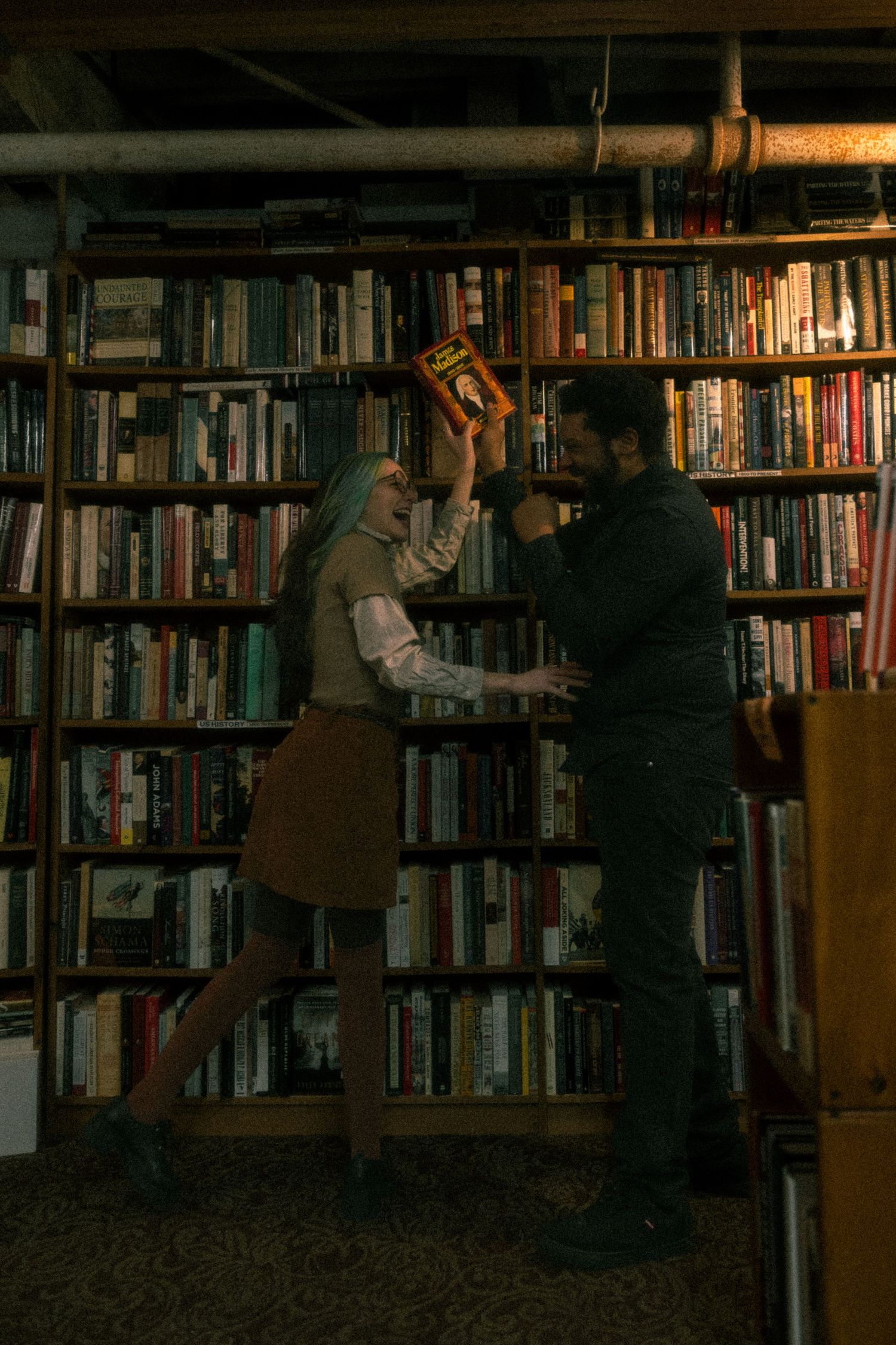 Two people browse books together in a dimly lit library with tall wooden bookshelves filled with literature.