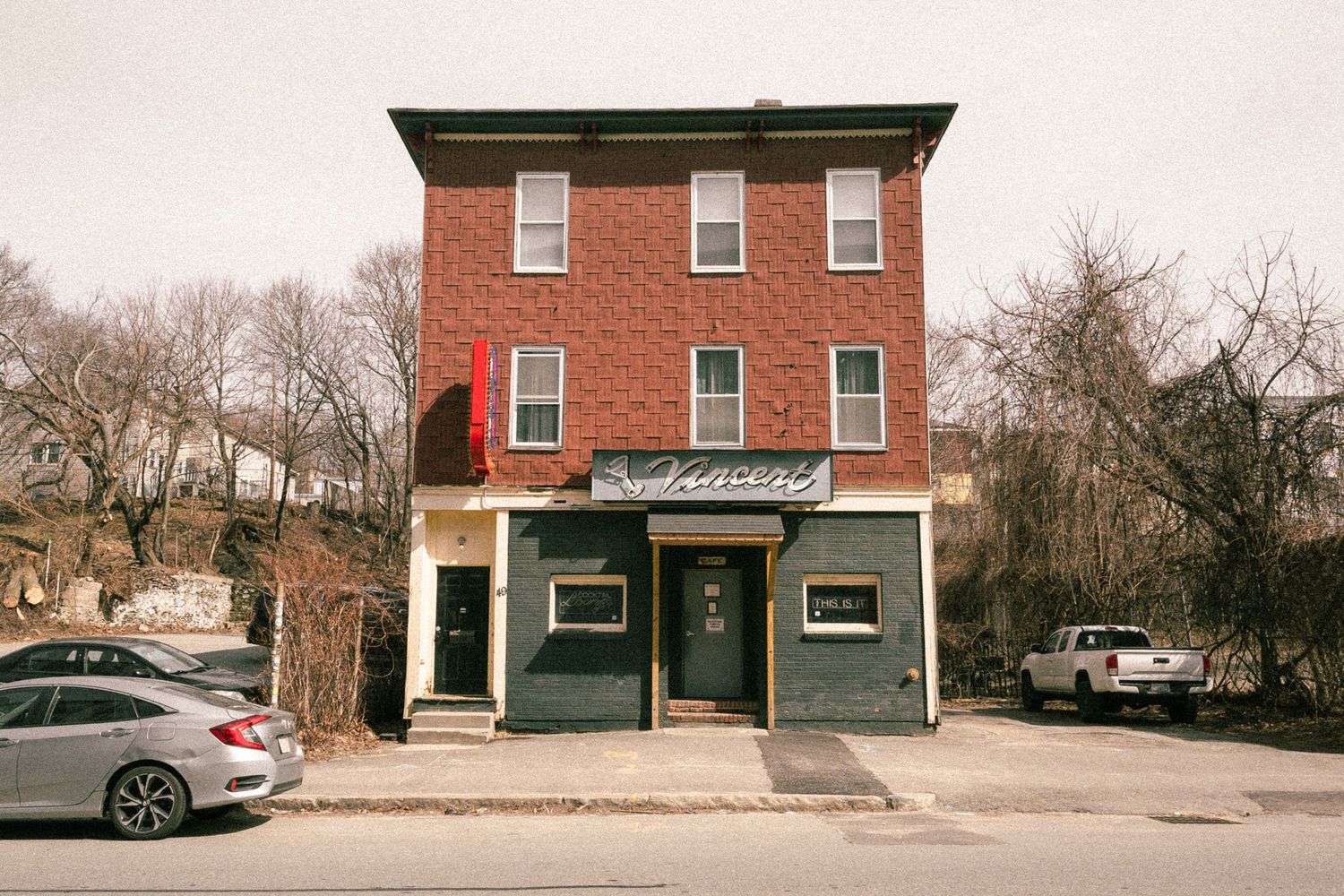 A red brick three-story building housing a bar on a quiet street corner with bare trees and parked cars nearby.