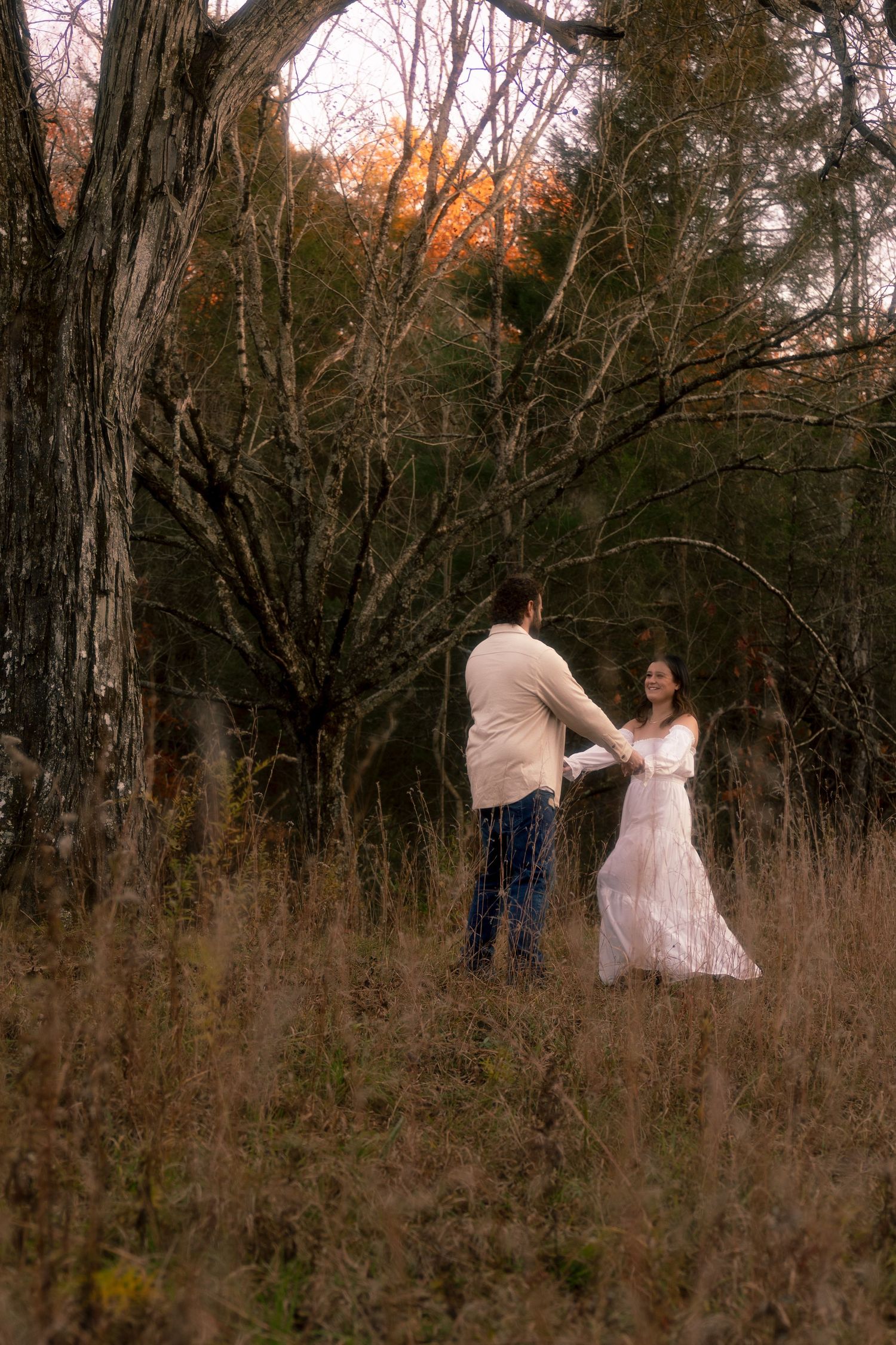 A couple dances together in a rustic field with bare trees and tall grass at sunset.