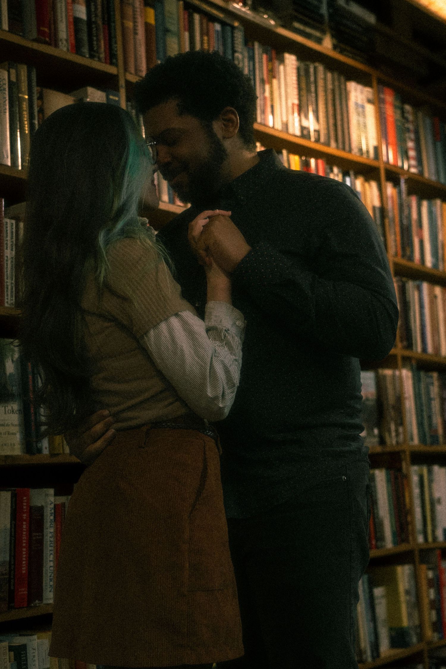 A romantic couple shares an intimate moment between bookshelves in a dimly lit library or bookstore.