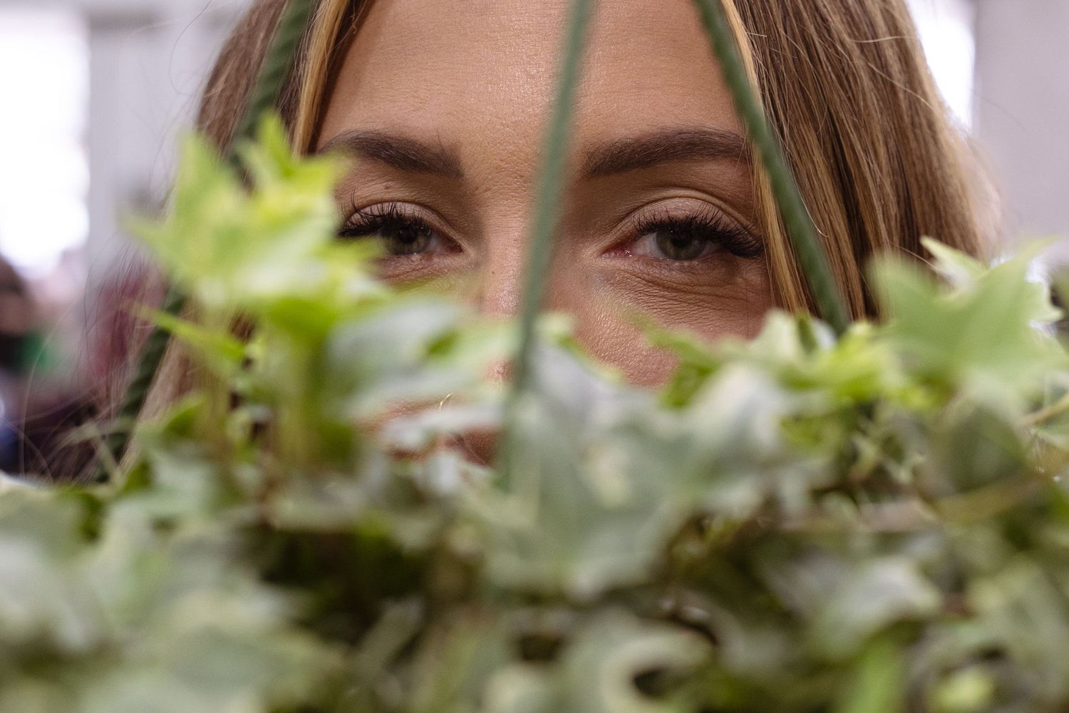 A person smiles behind fresh green herbs growing in a garden or planter.