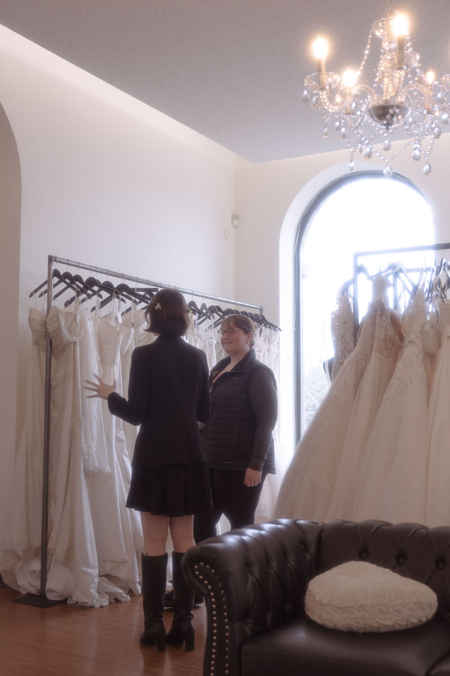 Two women browsing white wedding dresses on a rack in an elegant bridal boutique with chandelier lighting.