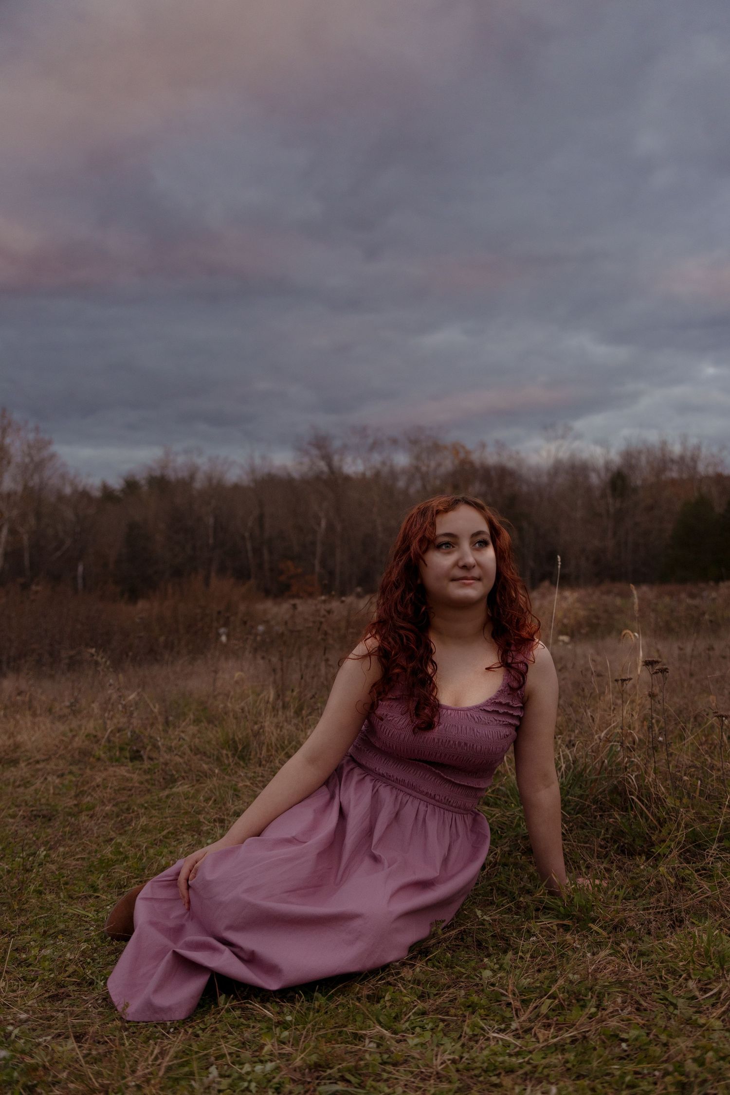 A woman in a mauve dress poses gracefully in a rustic field under moody autumn skies.