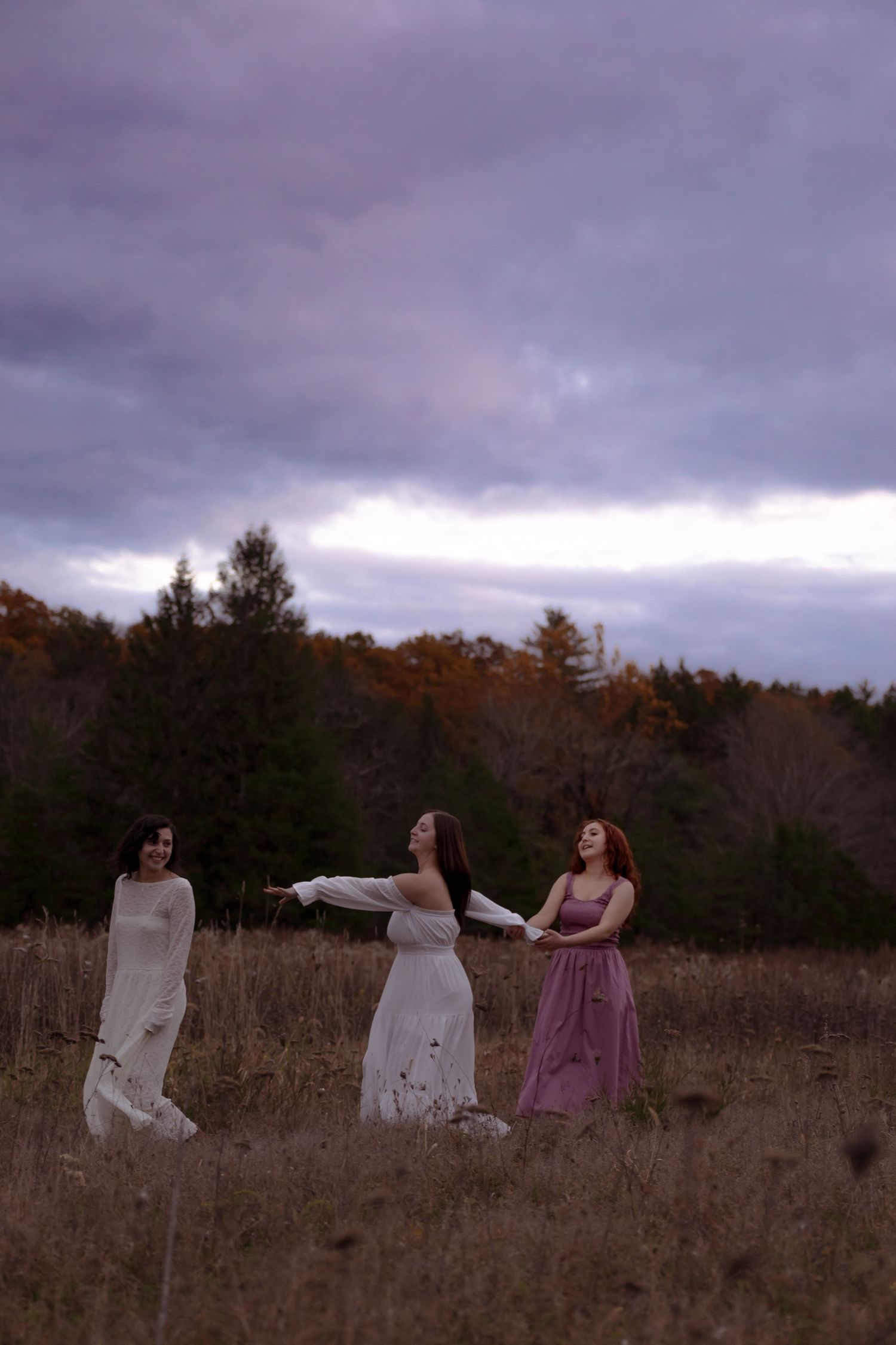 Three people in flowing dresses dance through a misty autumn field at dusk under dramatic purple clouds.
