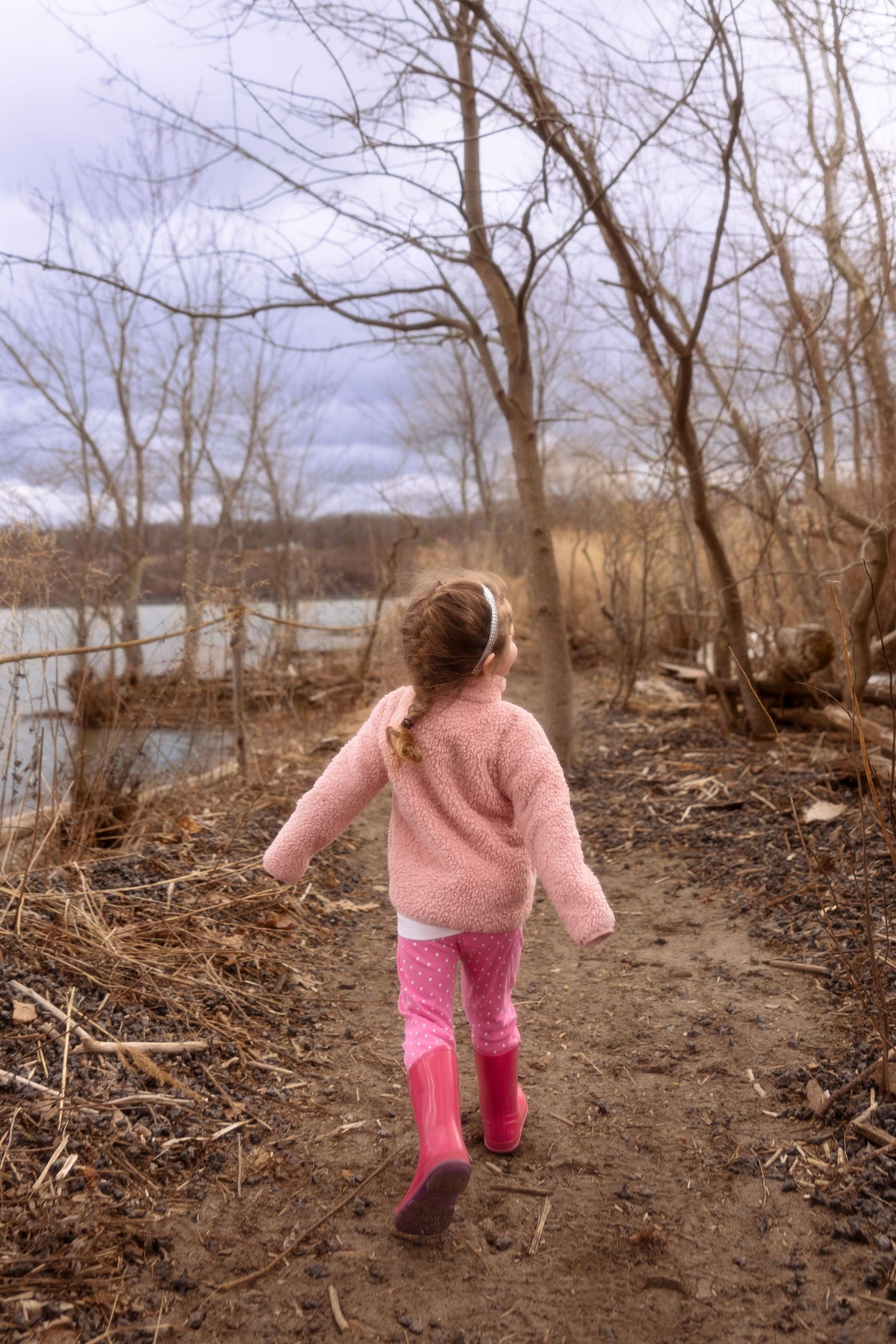 A child in a pink sweater and rain boots walks along a muddy trail by a lake on a cloudy winter day.