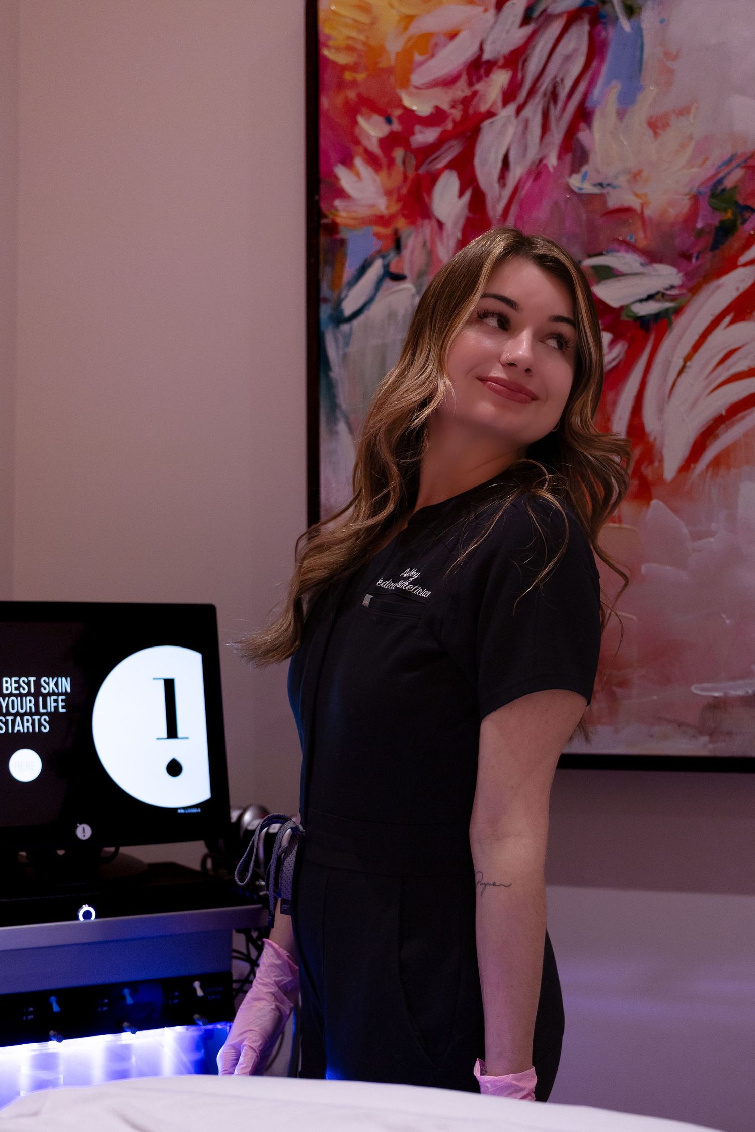 A person in black attire poses playfully against a backdrop of vibrant abstract artwork in a gallery setting.