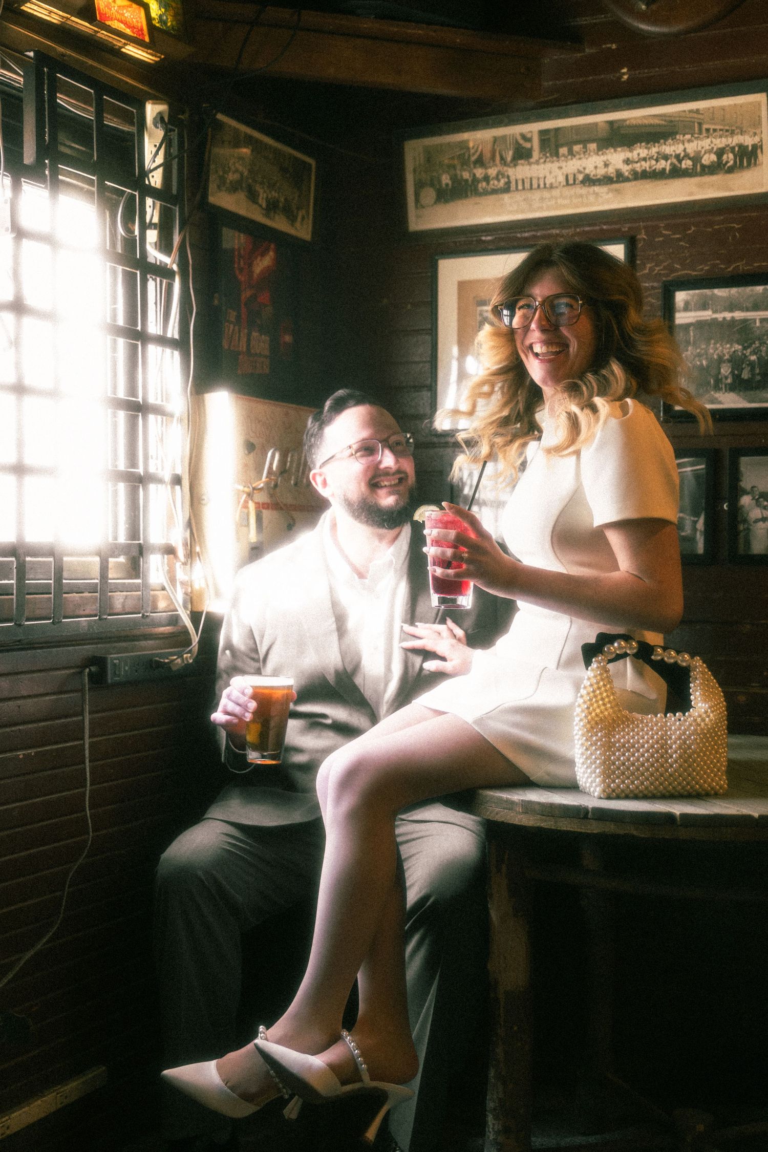Couple smiling and holding drinks in a cozy, warmly lit vintage bar setting.
