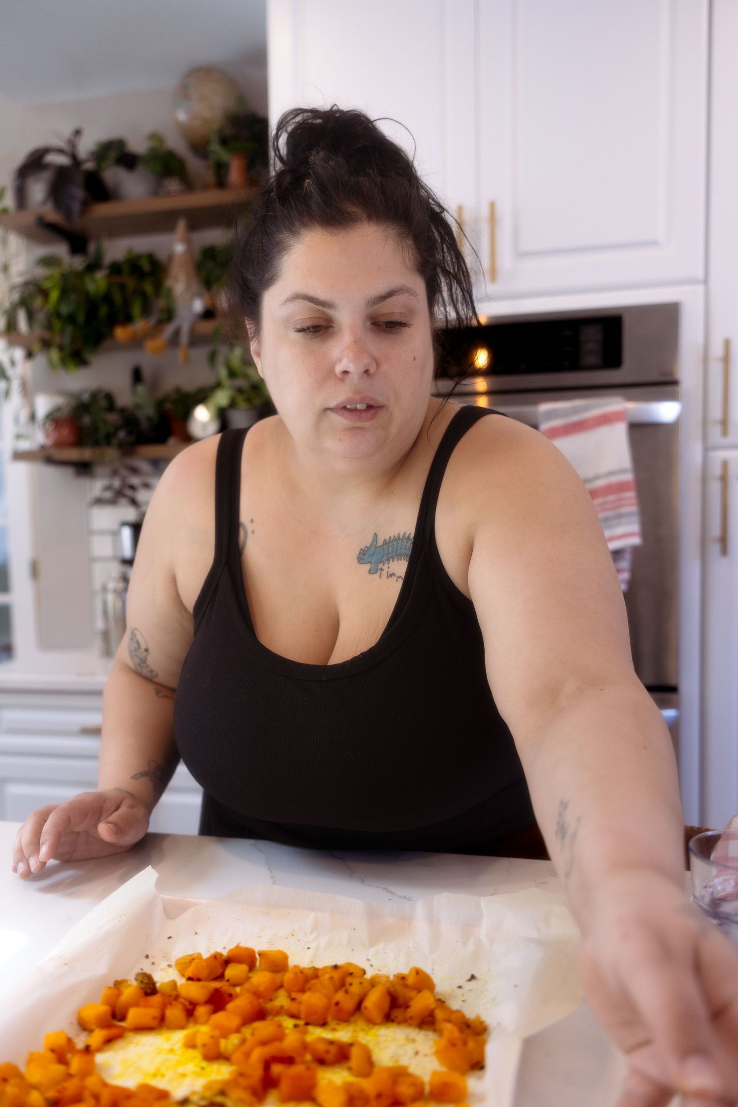 Someone in a black tank top prepares a colorful meal with roasted carrots in a bright modern kitchen with plants.