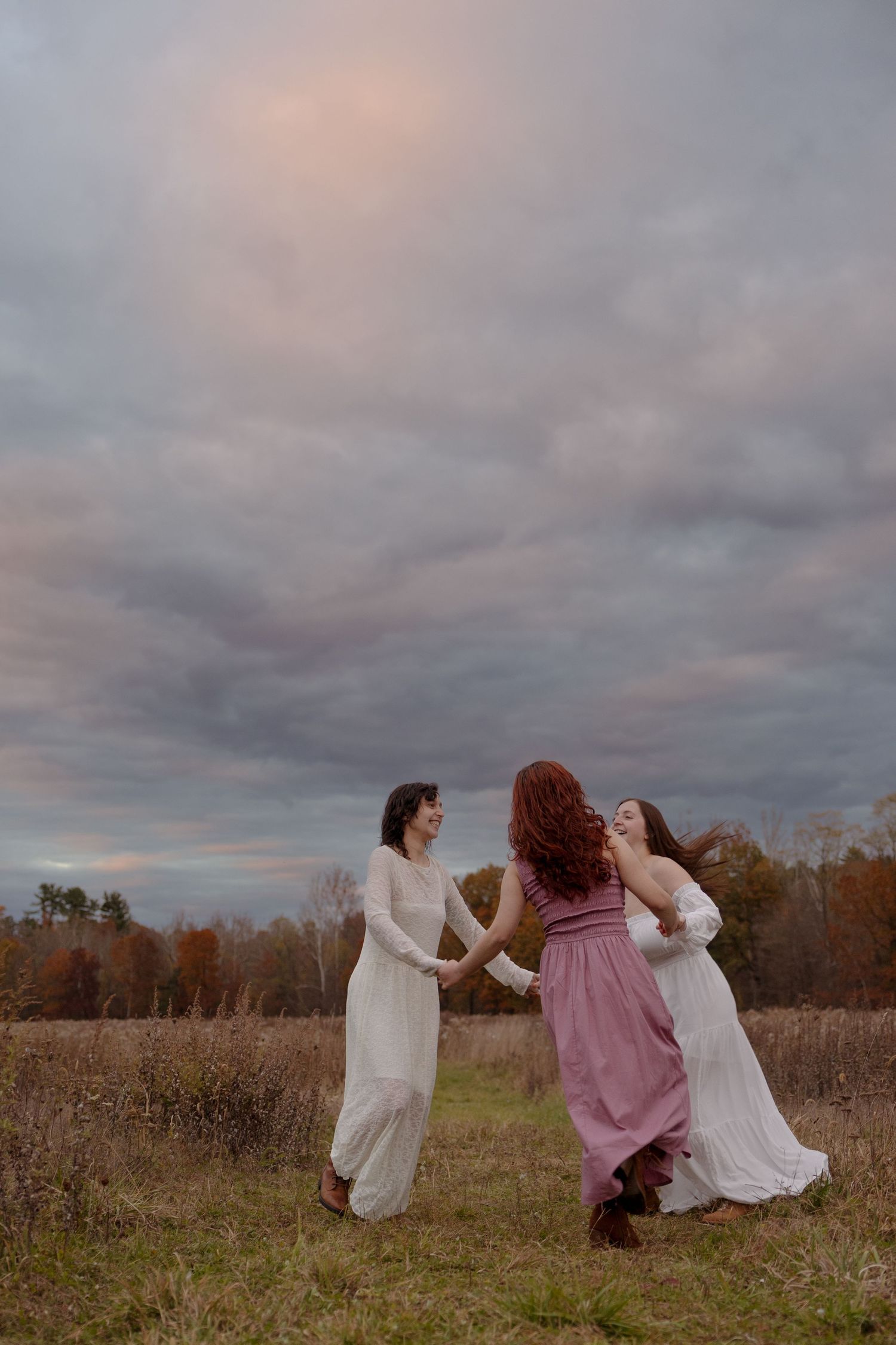 Three figures in flowing dresses dance and twirl together in a golden autumn field at dusk.