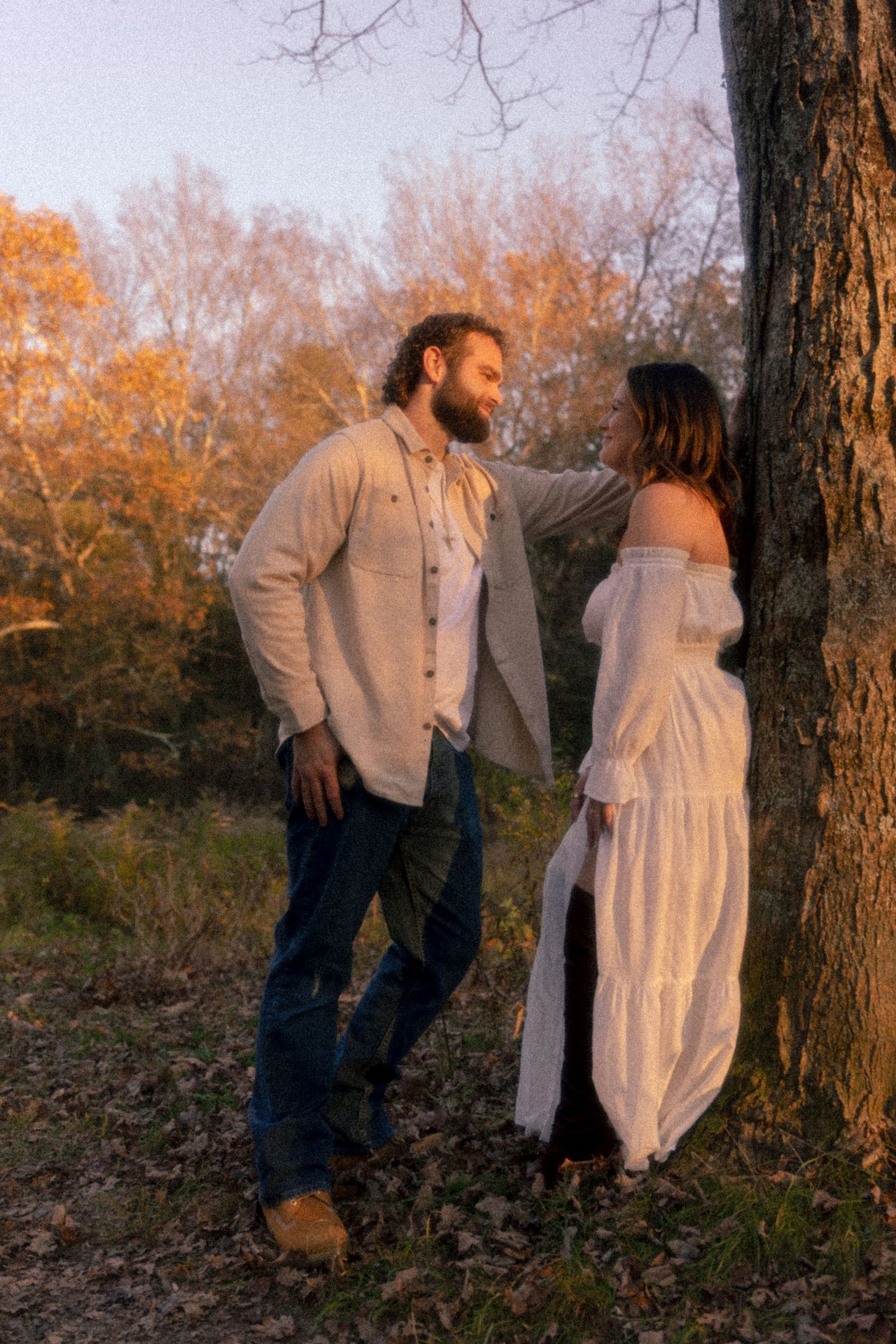 A couple in casual attire poses for romantic photos against autumn trees at sunset.