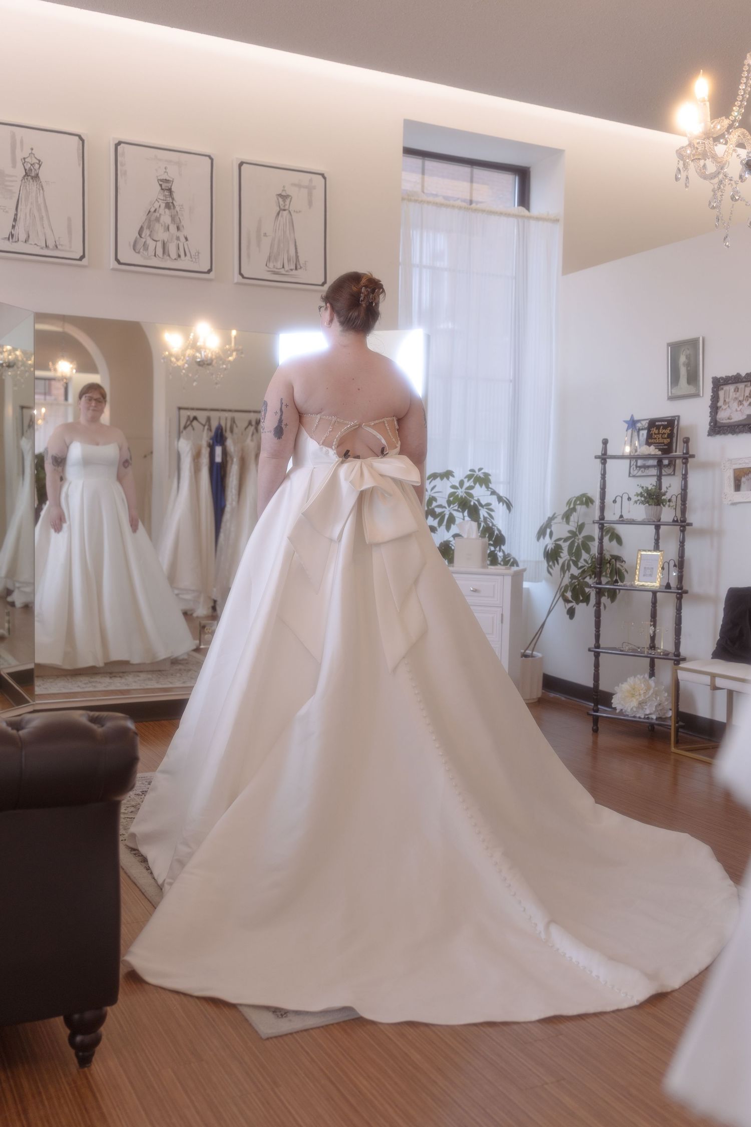 Bride in elegant backless wedding gown with large bow, long train, in bridal boutique with mirror reflection.