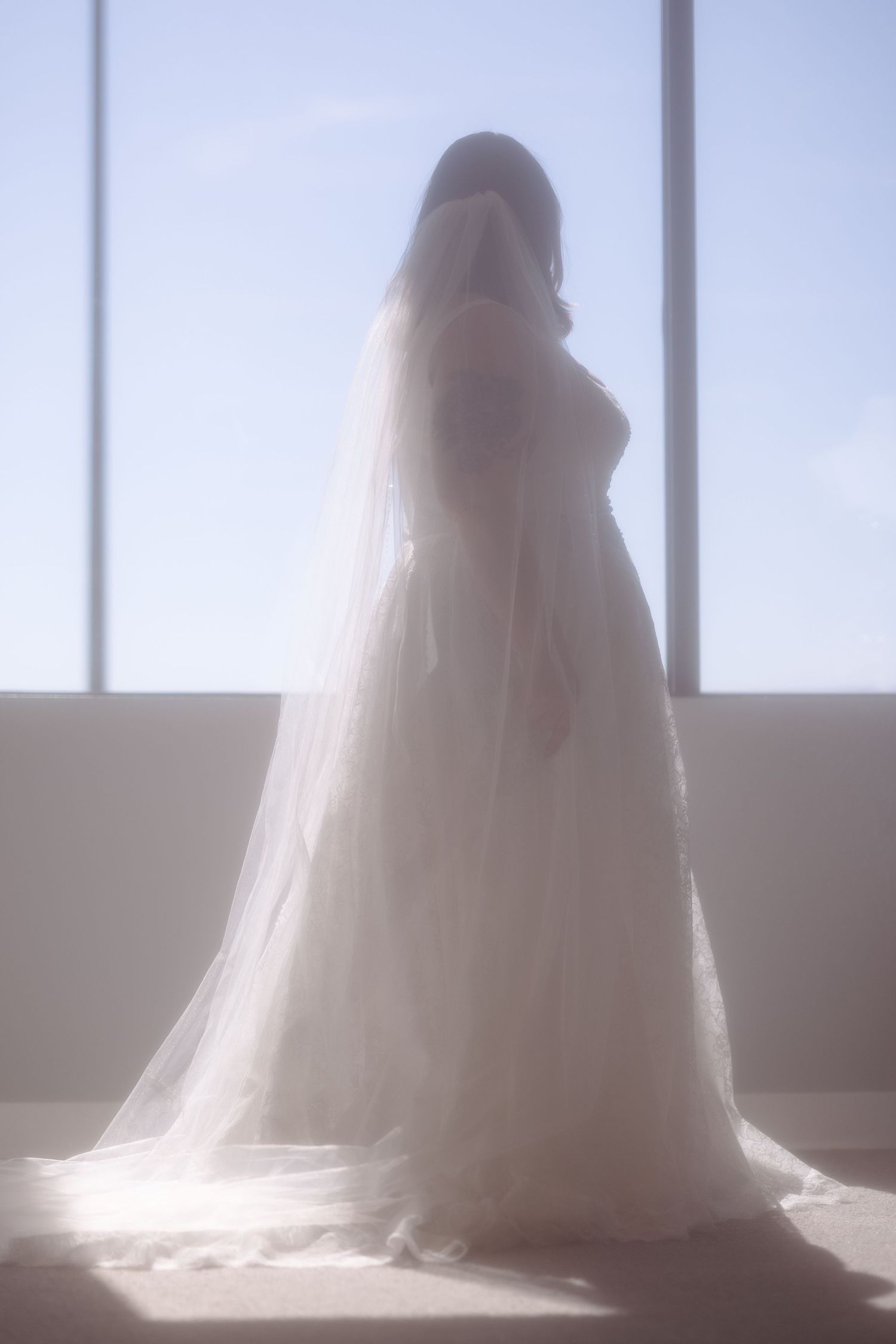 Bride in white wedding gown and veil standing backlit by large windows in soft ethereal light.