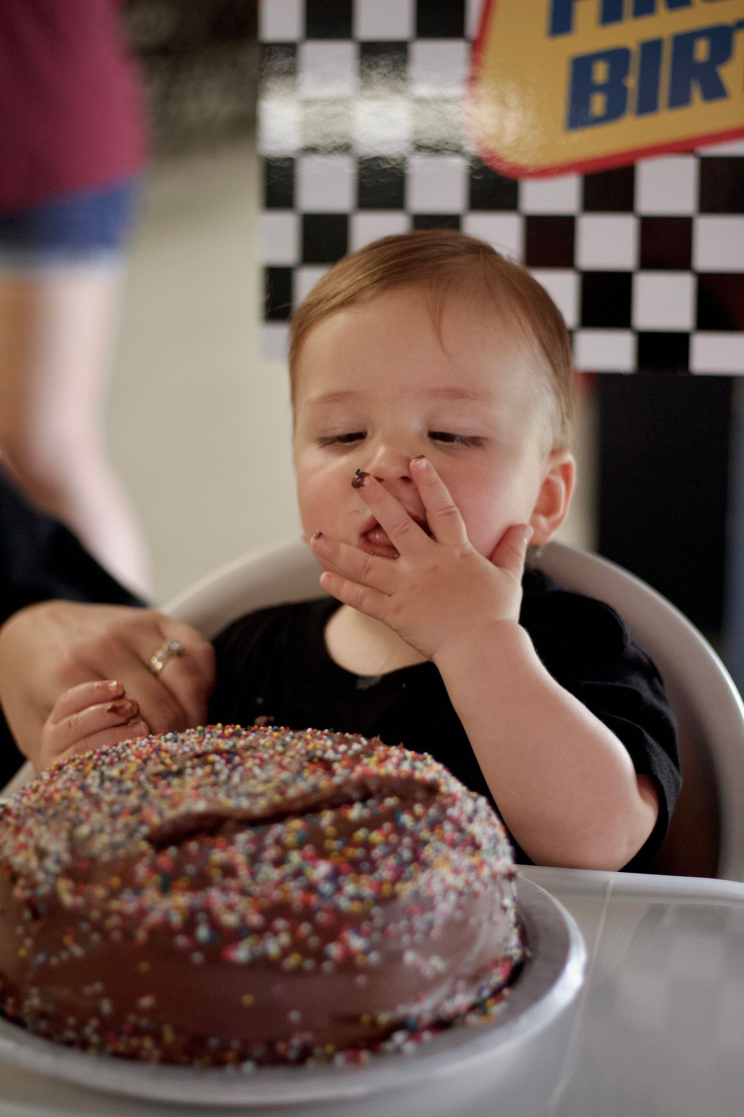 Baby giggles with chocolate-covered fingers near a sprinkle-decorated birthday cake on checkered background.
