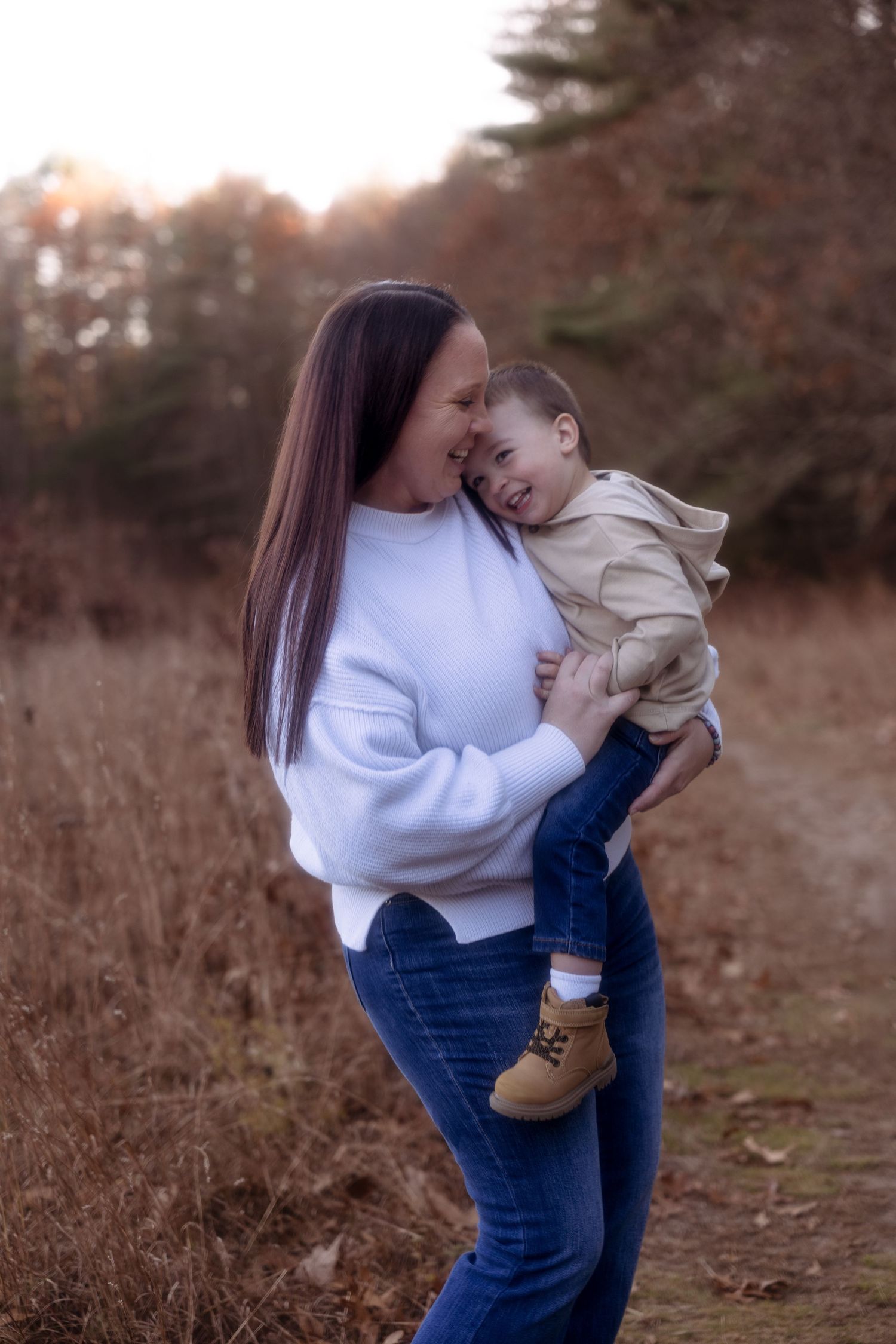 A person in a white sweater and jeans holds a small child wearing brown boots in an outdoor autumn setting.