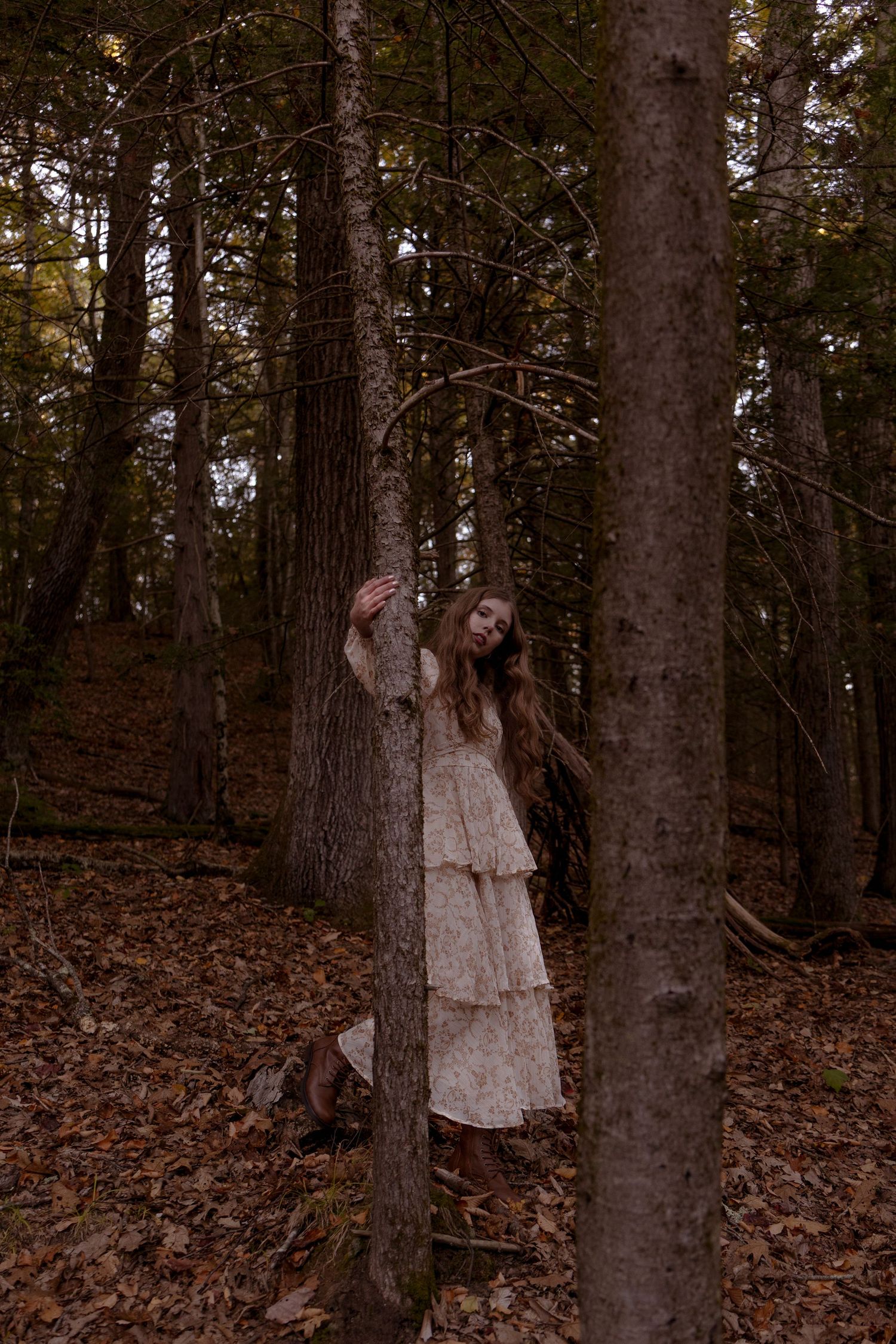 A figure in a vintage-style dress stands mysteriously among tall trees in a dark autumn forest setting.