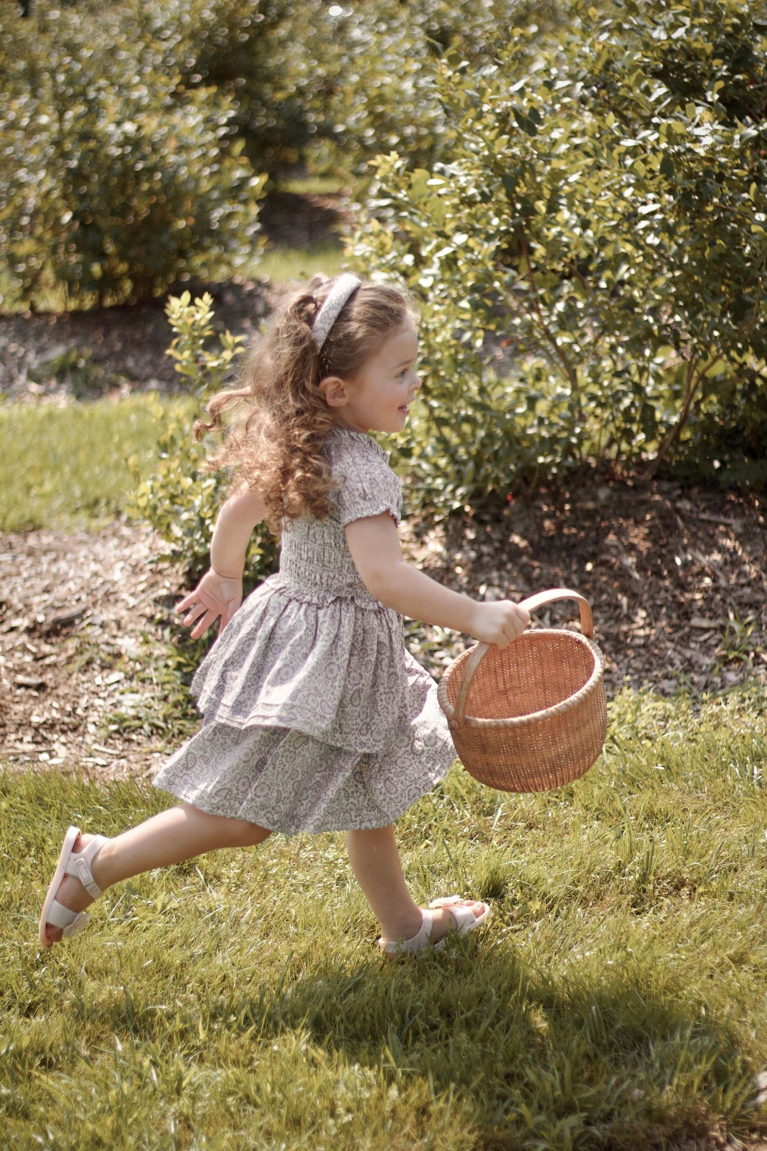 A child in a floral dress runs through a sunny garden carrying a wicker basket, creating a whimsical spring scene.