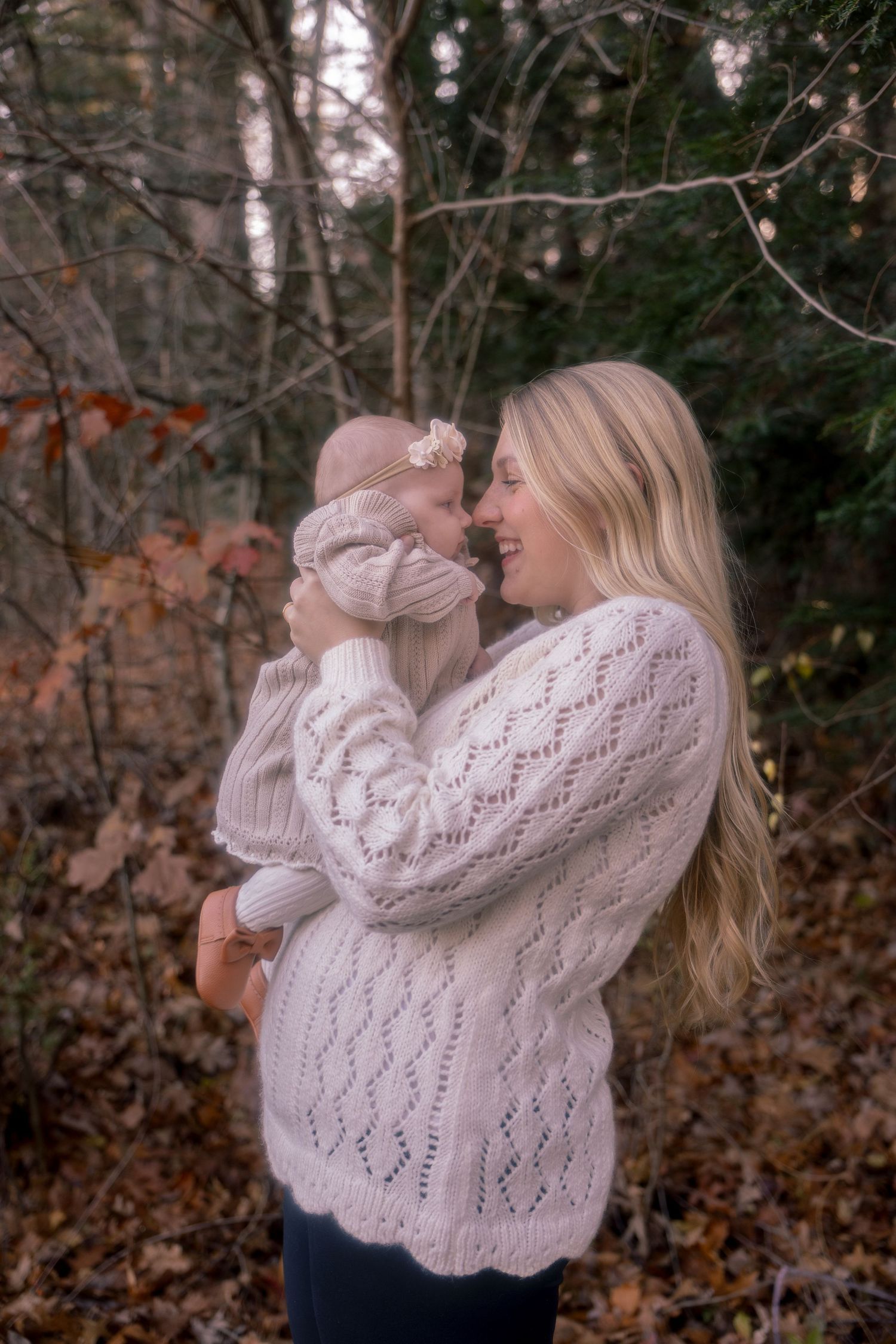 A parent and baby wearing matching white knit sweaters embrace outdoors among autumn trees.