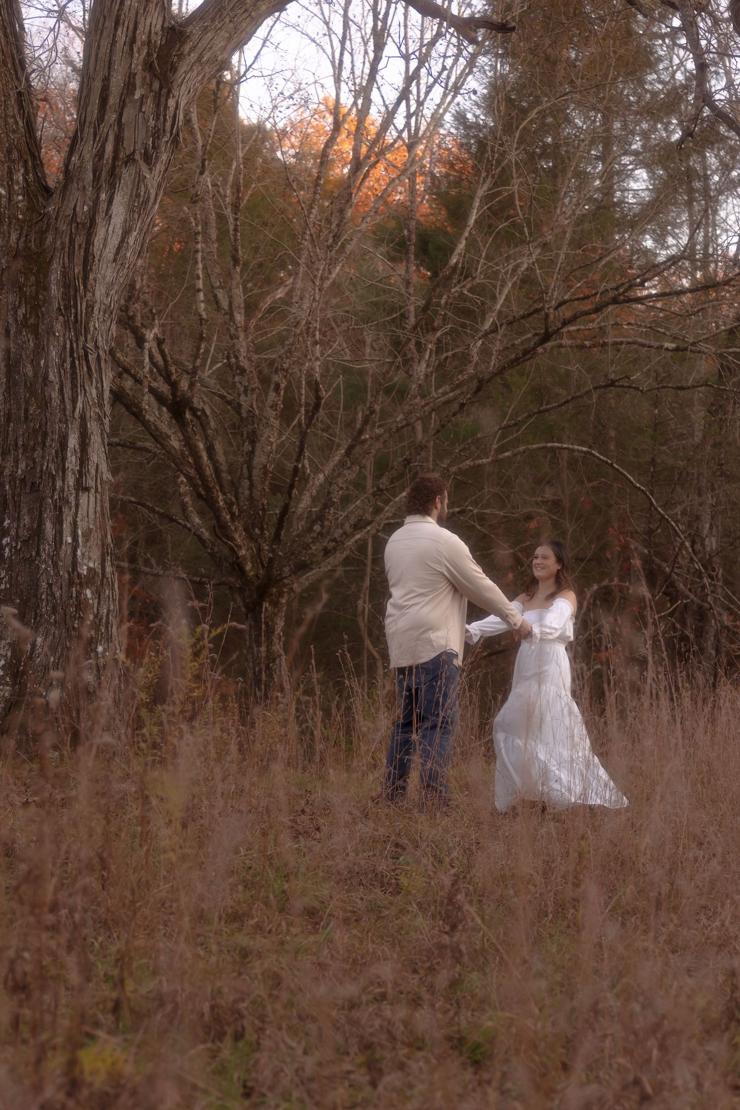 A couple dances together in a rustic outdoor setting with tall grass and bare trees during autumn.