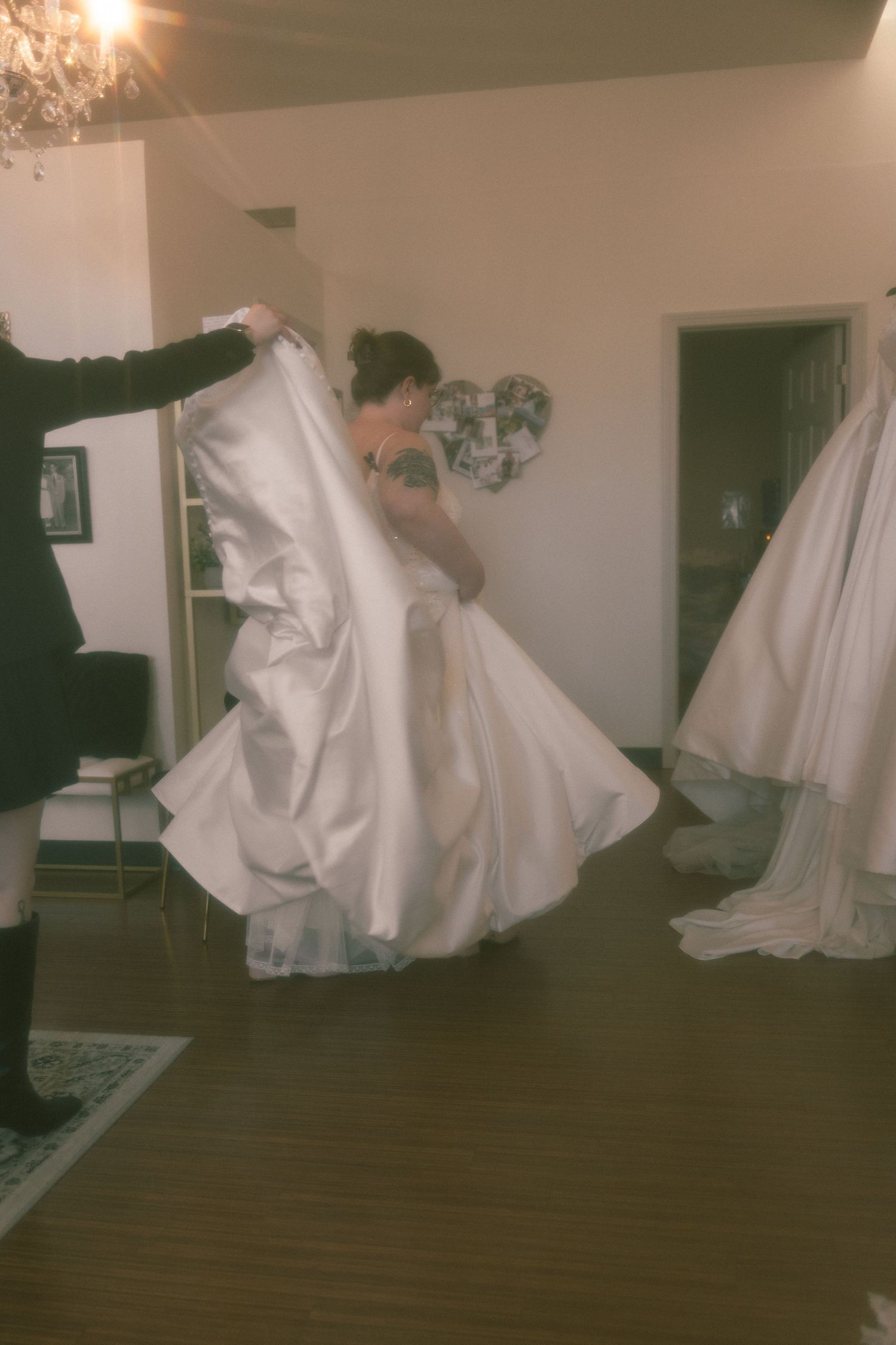 A bride in a white satin ball gown being helped with her dress in a softly lit room.