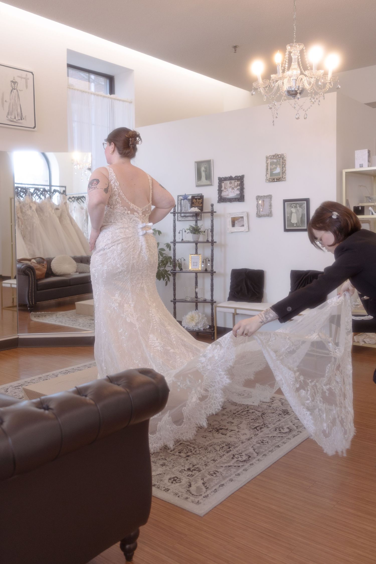 Bridal consultant adjusting lace wedding dress train on bride in elegant bridal boutique with chandelier.