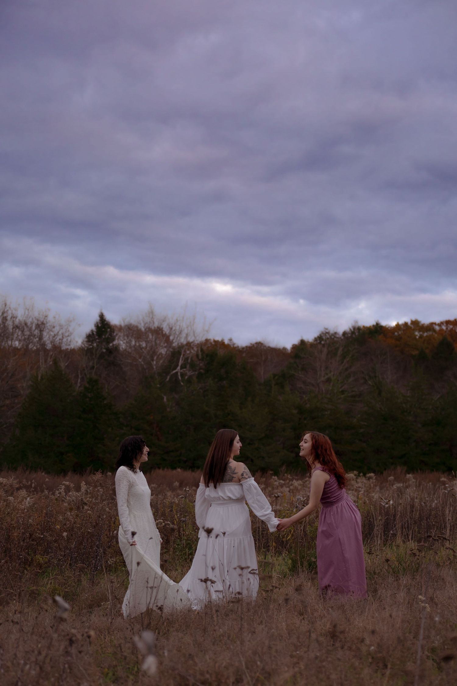 Three people in flowing dresses stand together in a moody autumn field under cloudy skies at dusk.