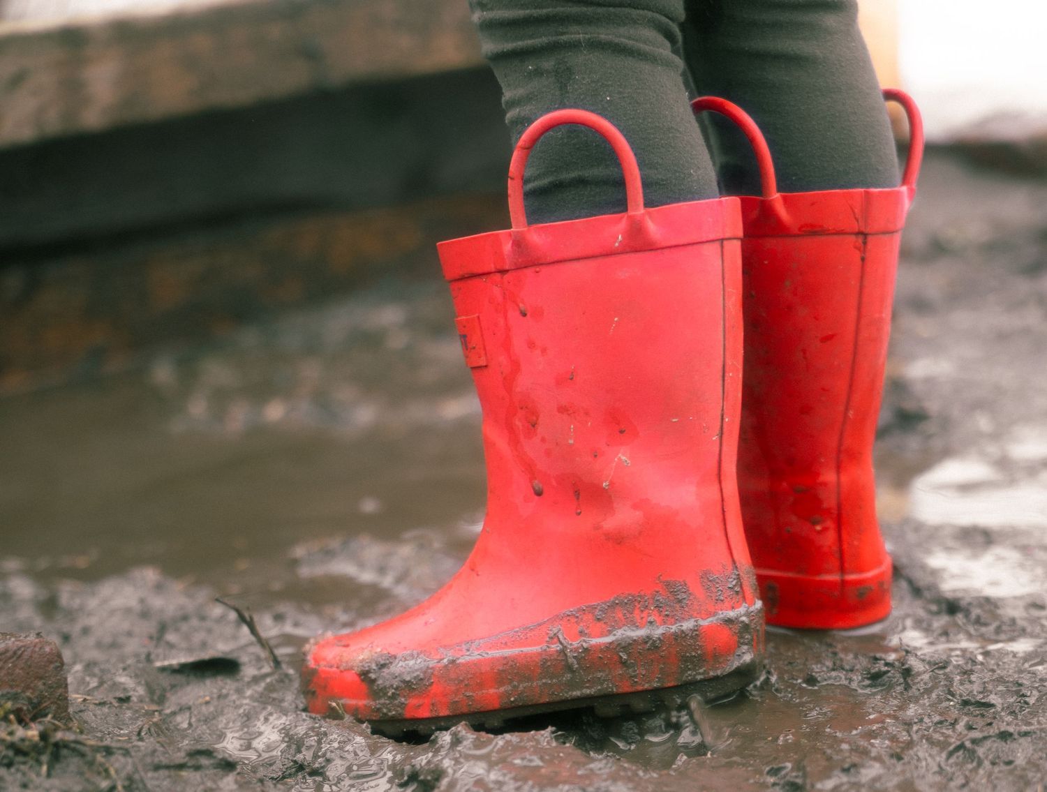 A pair of bright red rubber rain boots splashing through wet puddles on a rainy day.