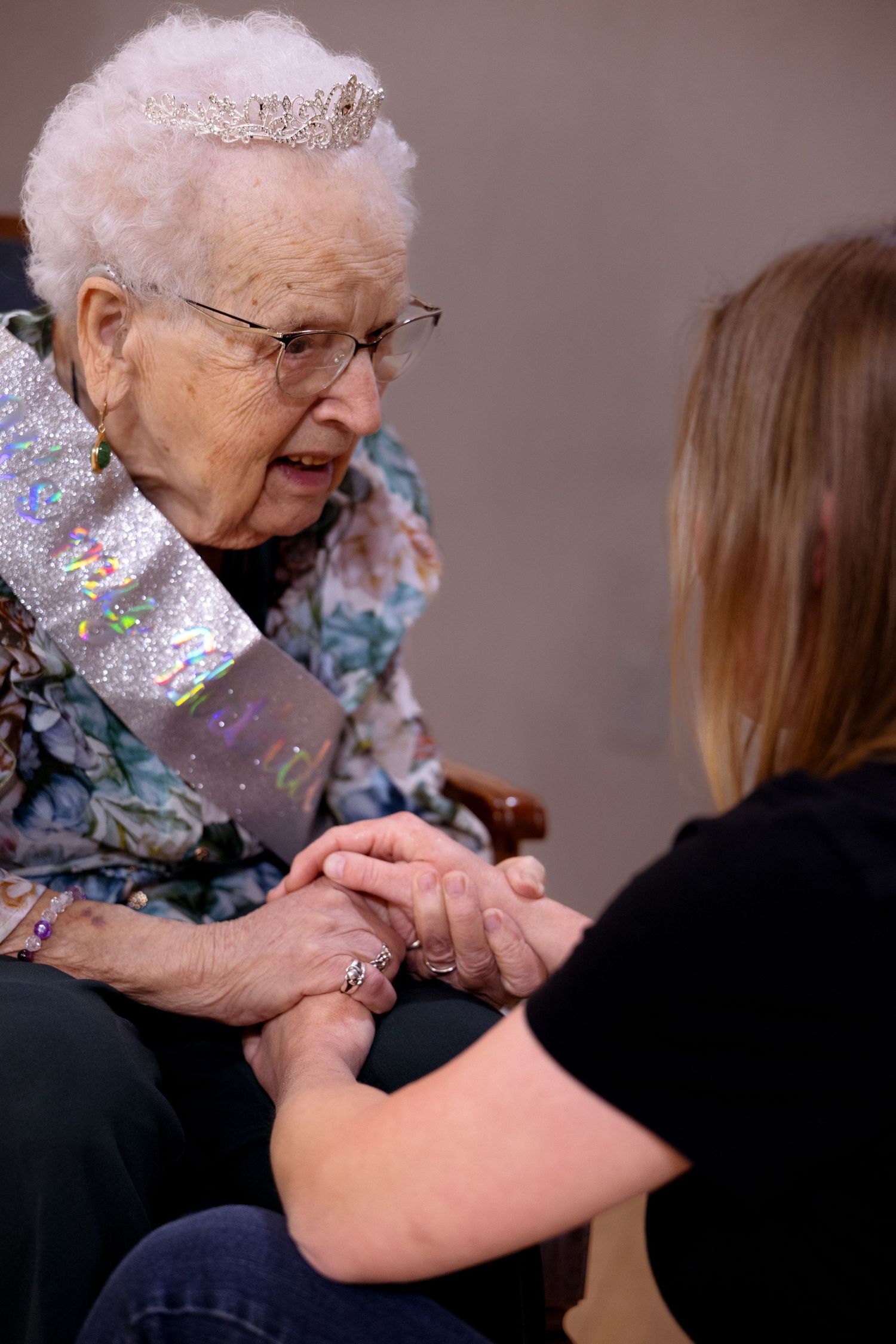 An elderly person wearing glasses and a floral scarf shares a tender moment with someone in a black shirt.