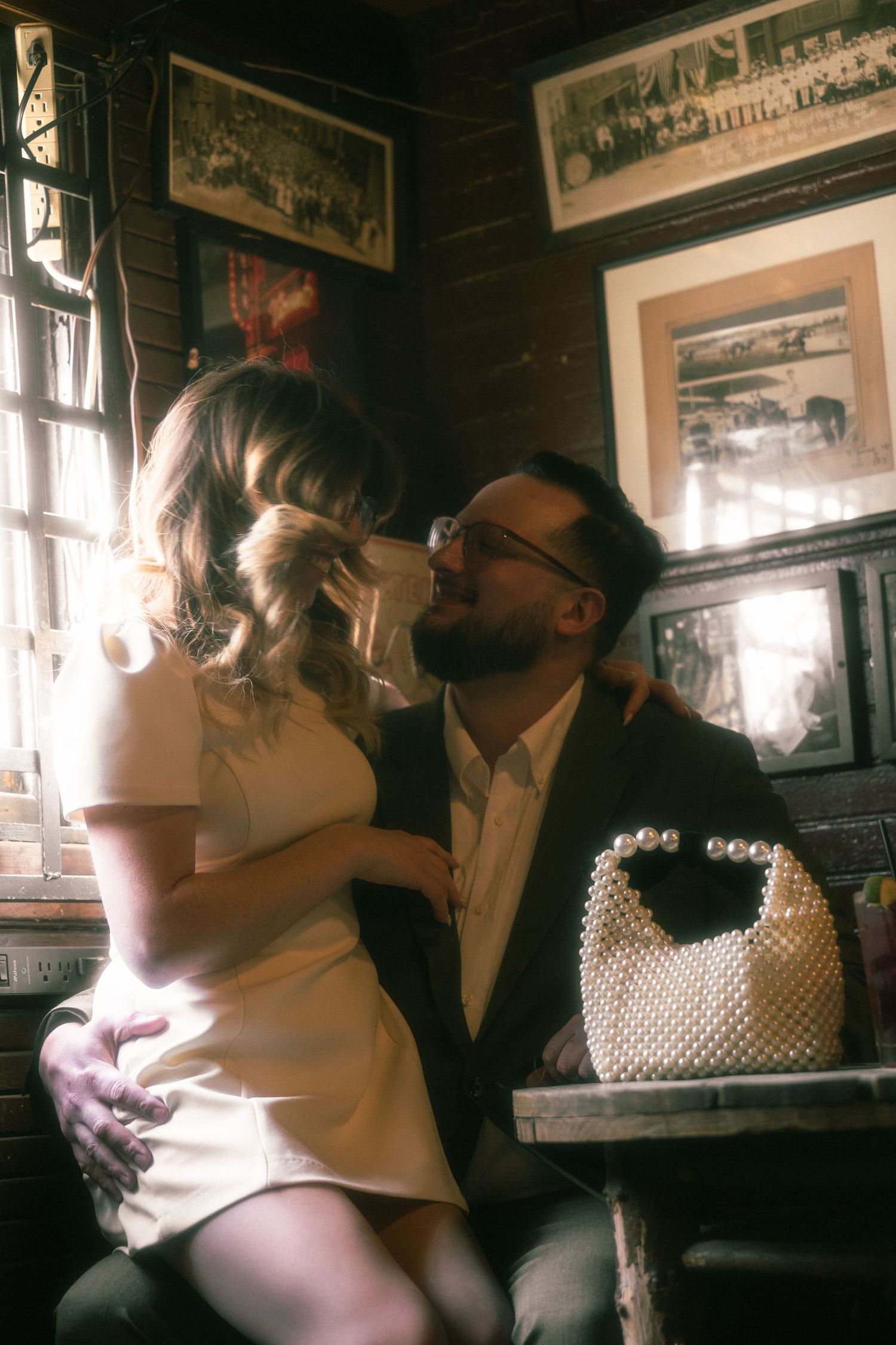 Couple sharing a romantic moment in a vintage bar, woman in white dress, man in suit with pearl handbag on table.