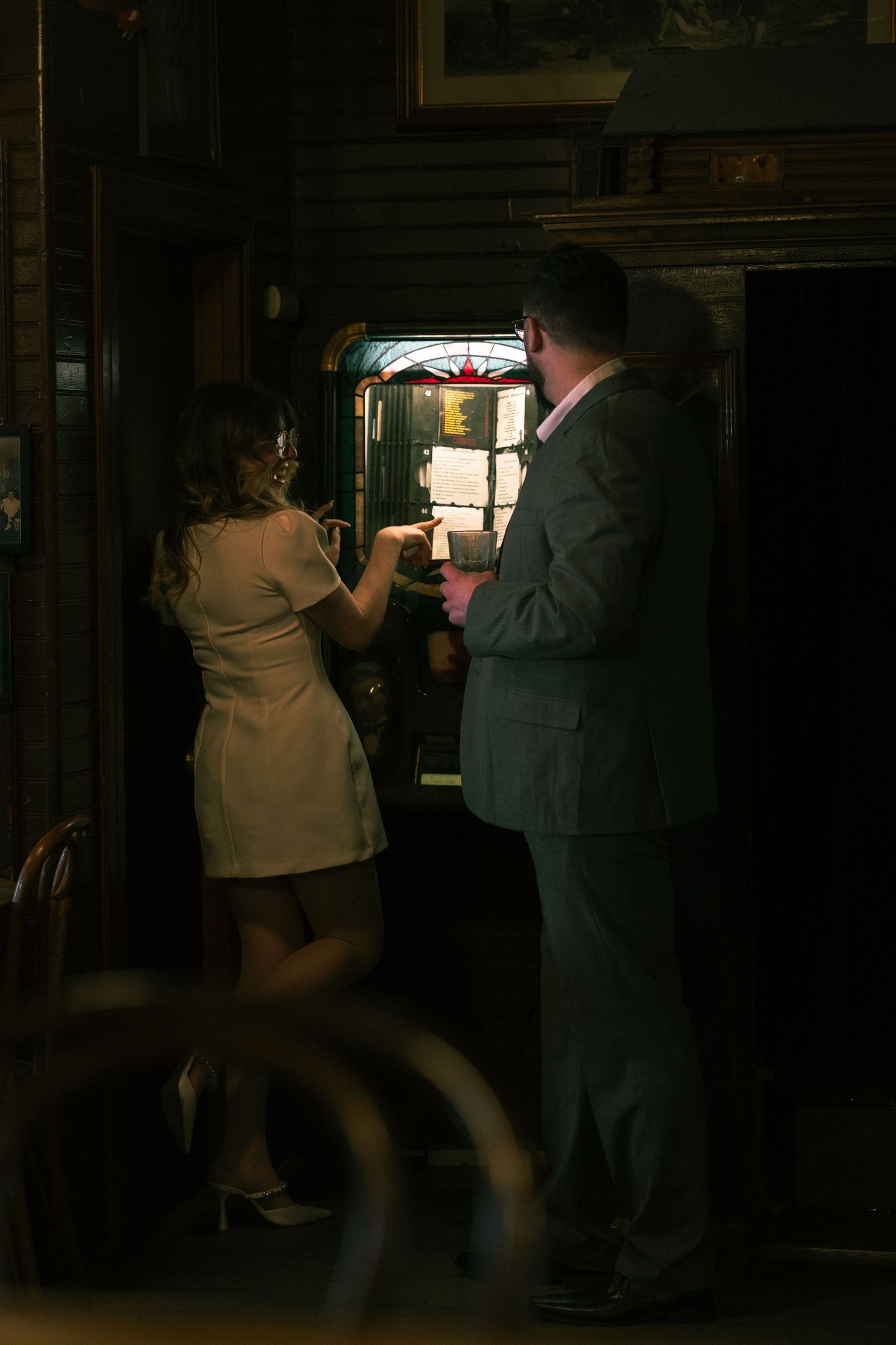 Couple standing by a glowing jukebox in a dimly lit bar, woman in white dress, man in gray suit.