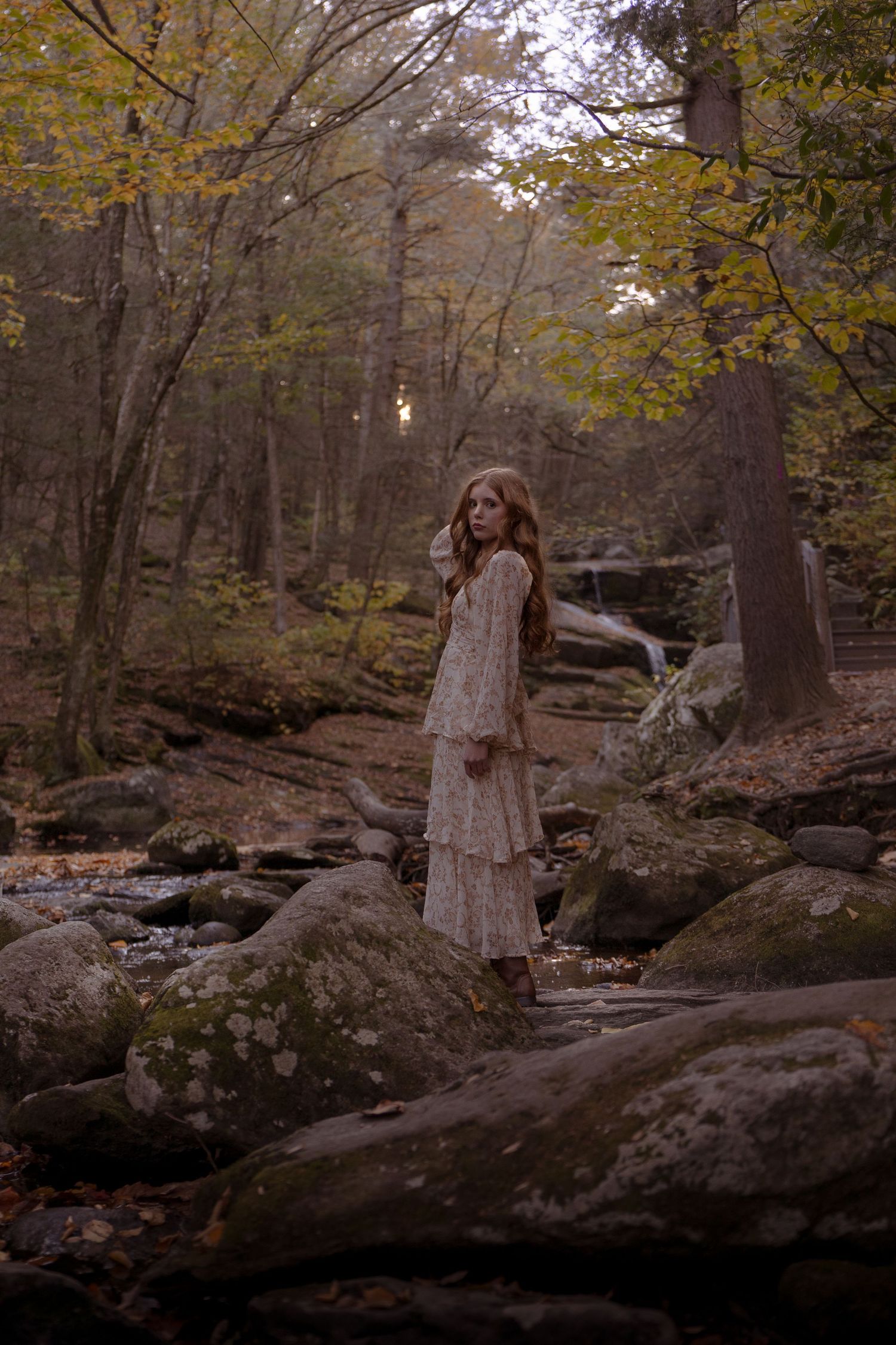 A figure in a vintage white dress stands on mossy rocks by a stream in an autumn forest.