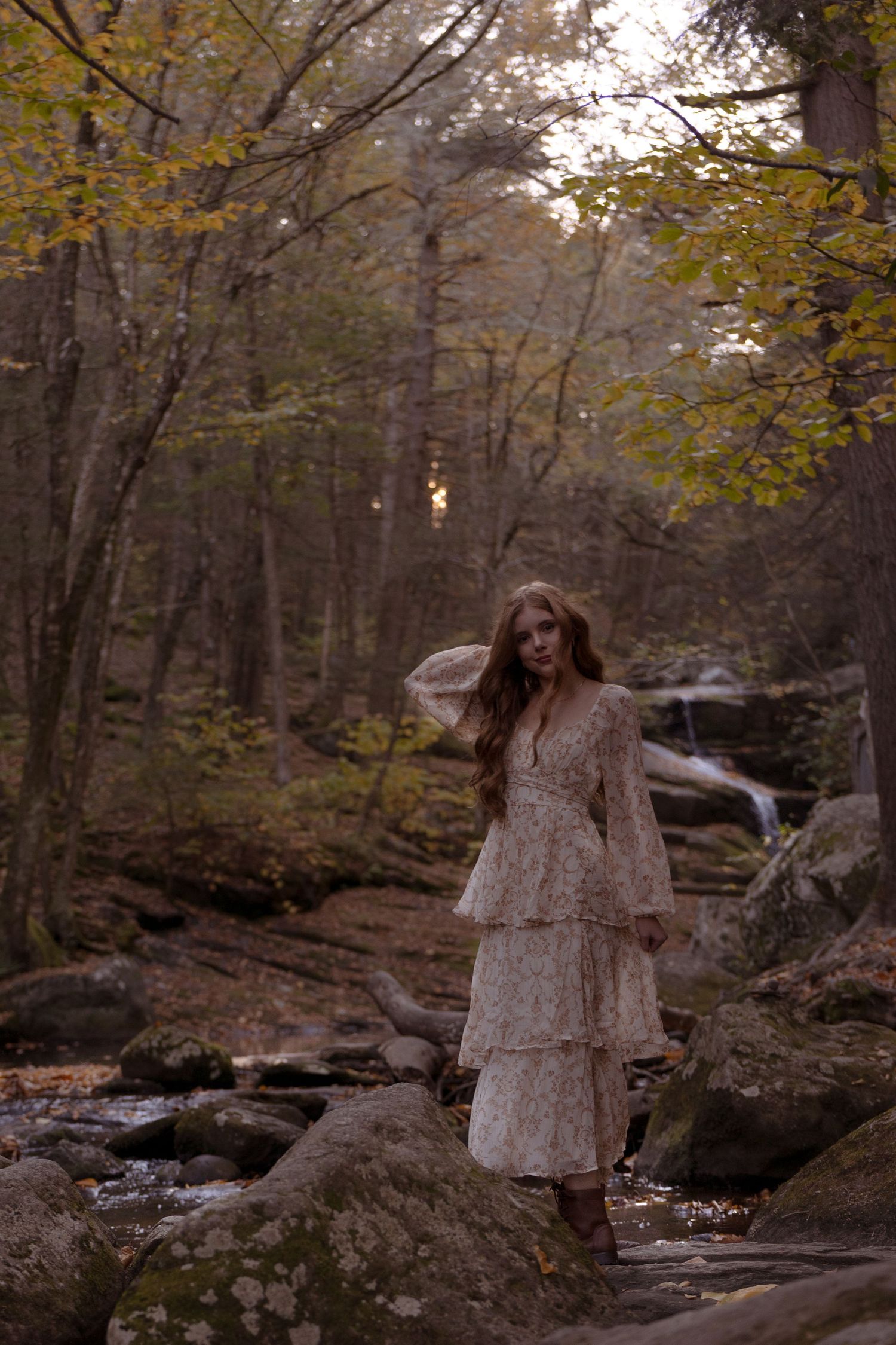 A figure in a flowing dress stands among autumn trees beside a rocky stream in a moody forest setting.