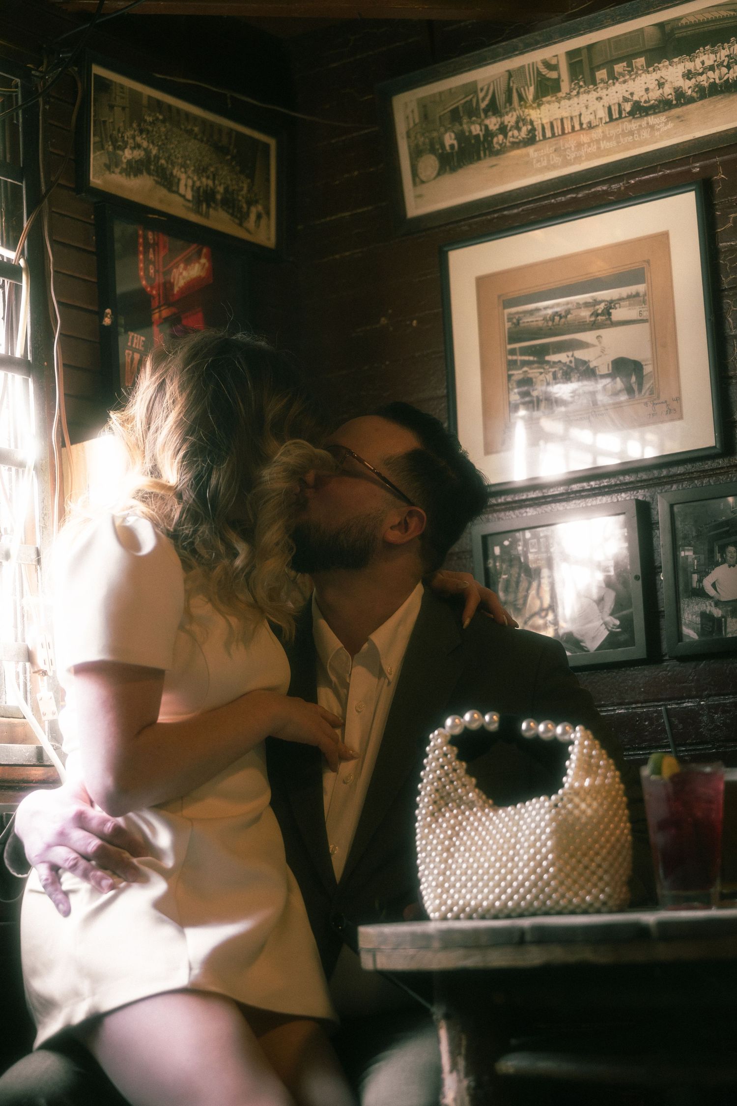 Couple sharing a romantic kiss in a cozy vintage bar, woman in white dress, man in suit, pearl handbag on table.