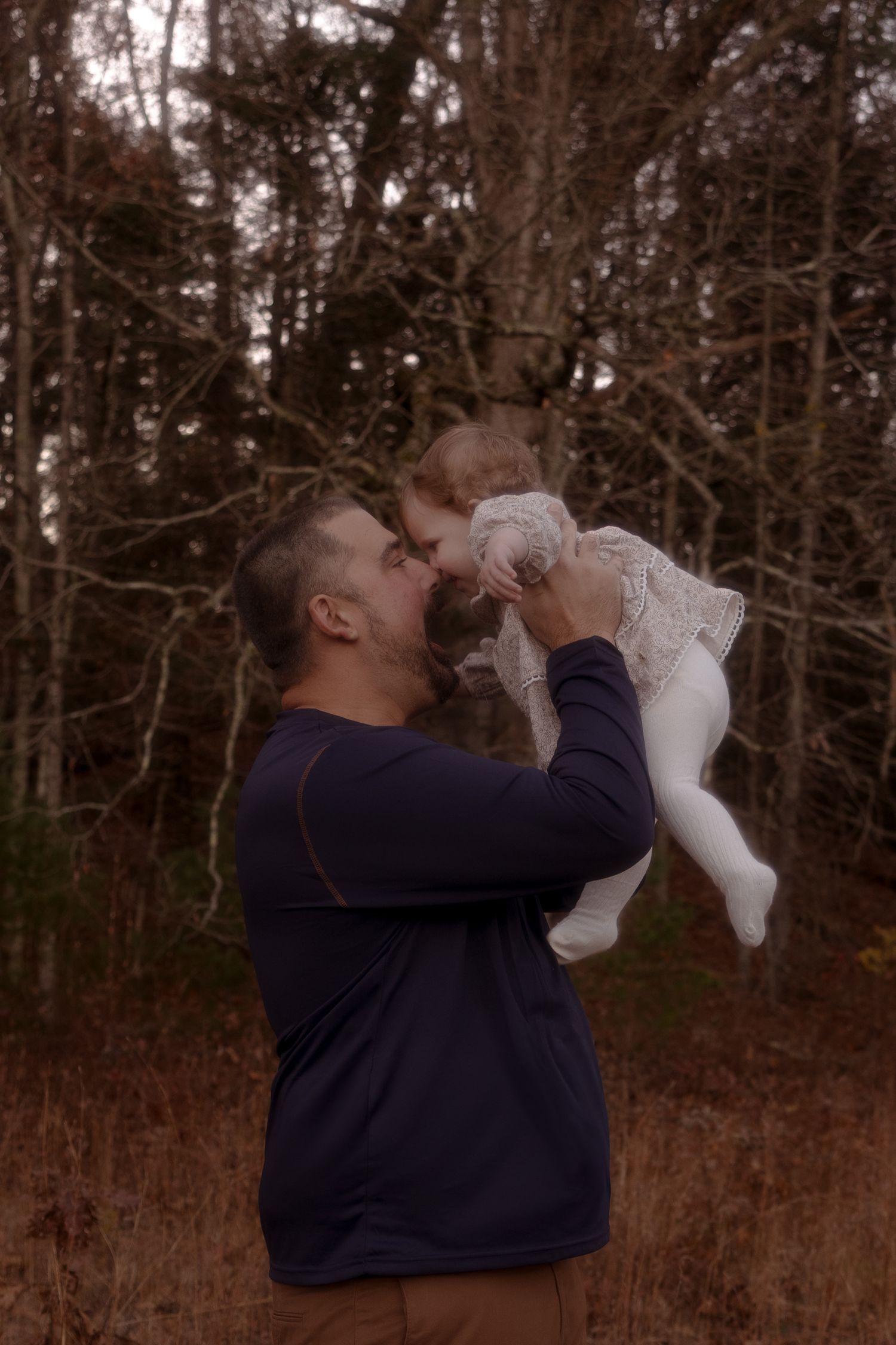 Person in navy sweatshirt holds and kisses small white dog in wooded outdoor setting.