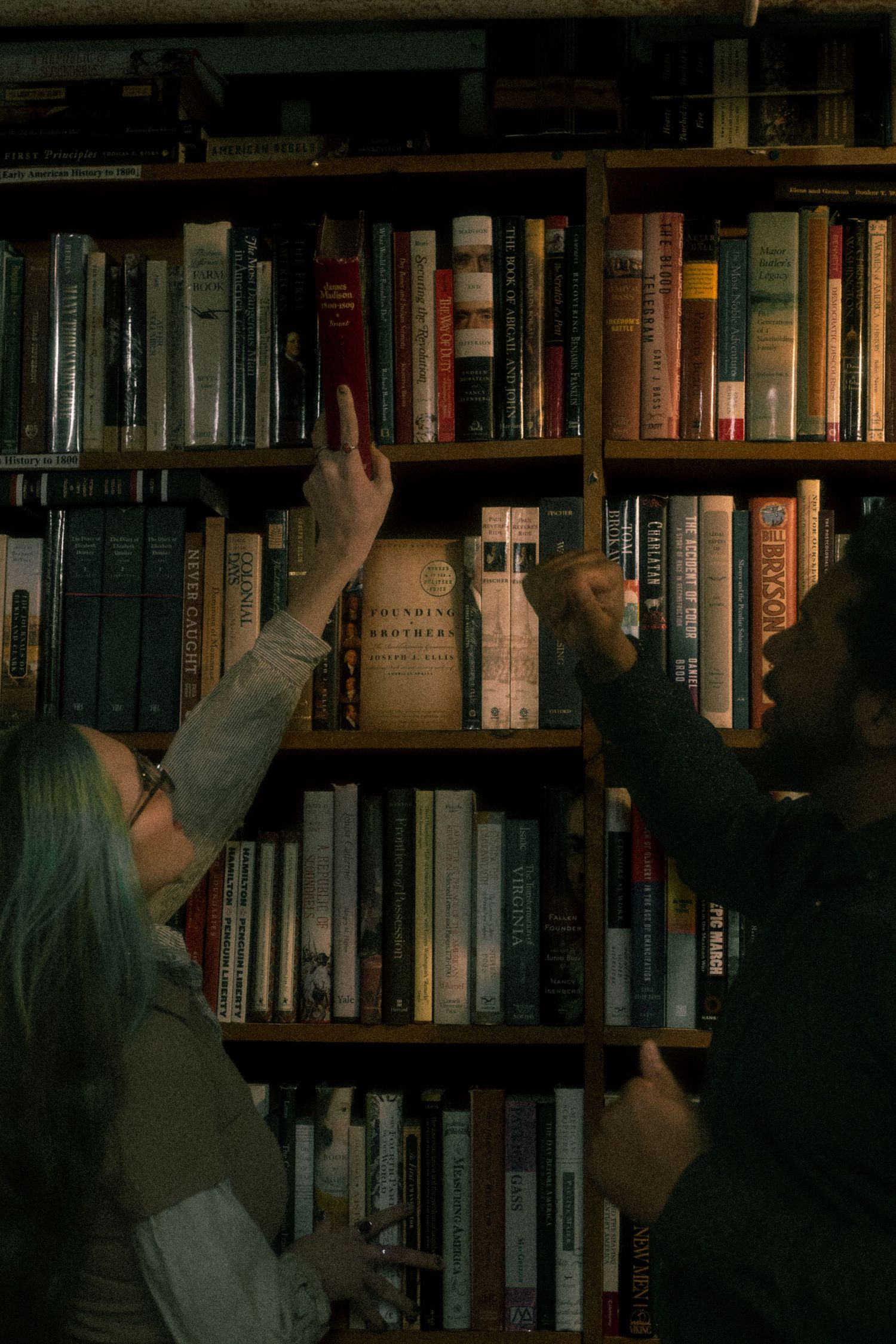 A person reaches up to grab books from tall wooden bookshelves filled with various hardcover volumes.