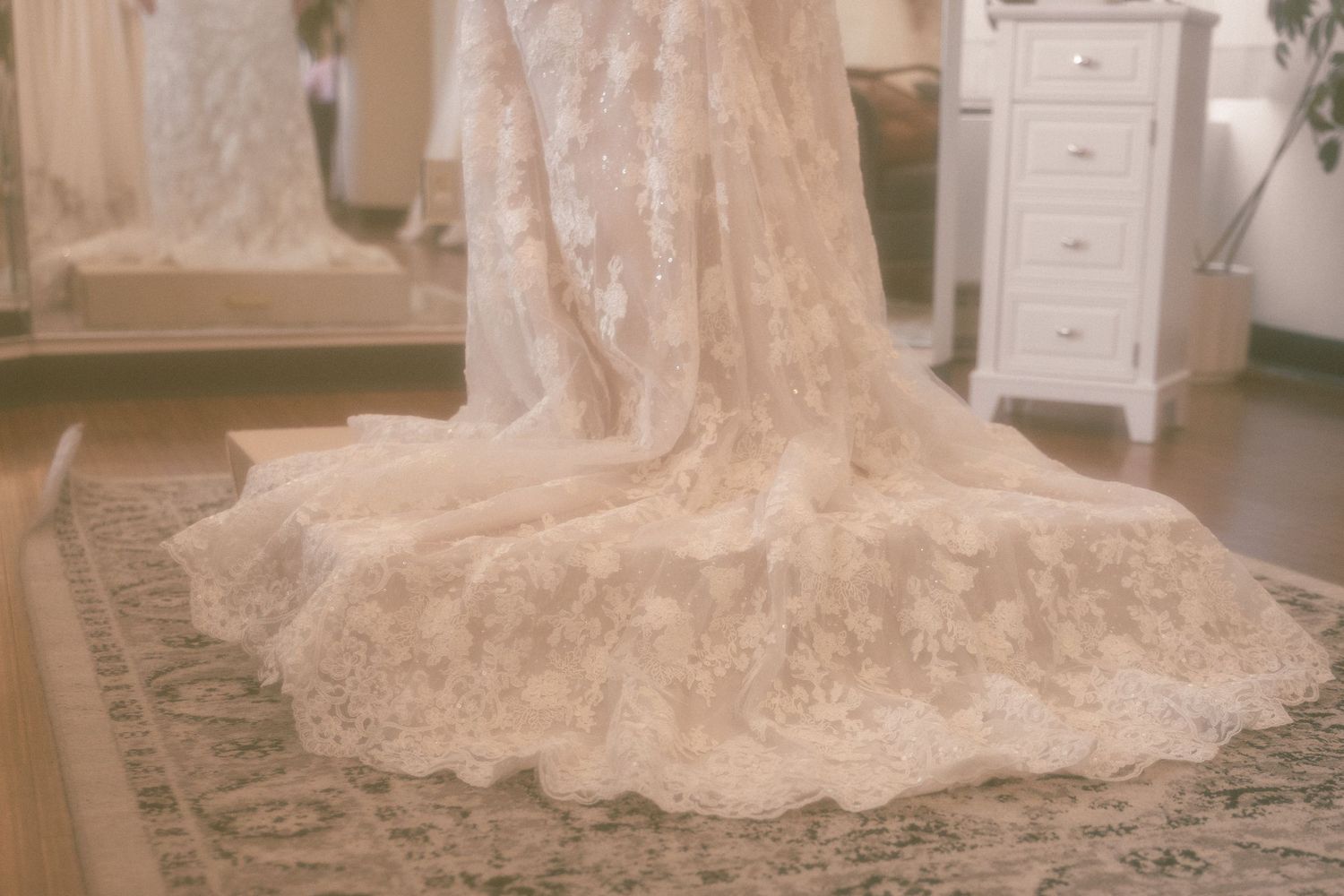 Elegant lace wedding dress train spread on a patterned rug in a bridal boutique fitting room.