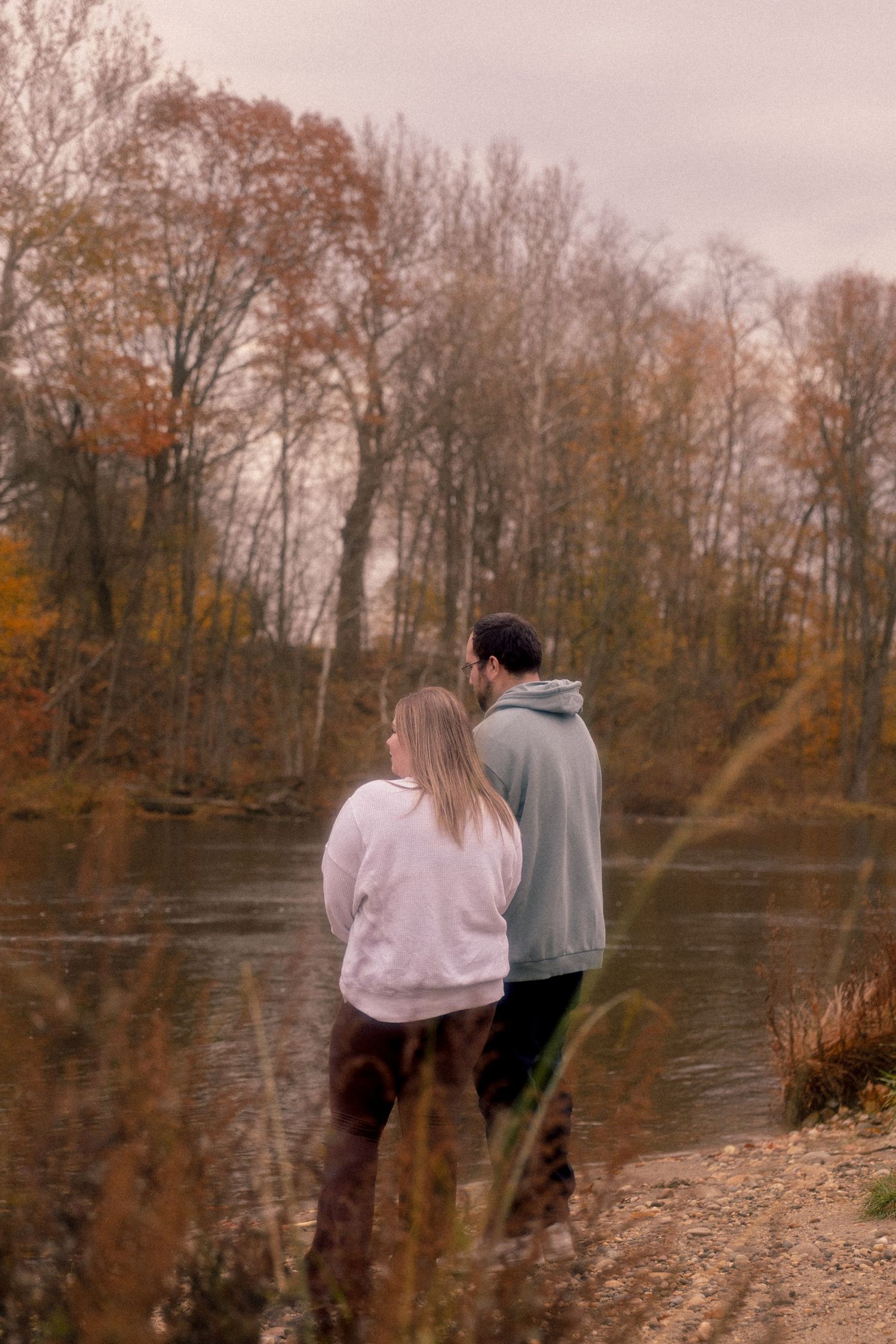 Two people stand at the edge of a serene lake surrounded by autumn trees on a cloudy day.