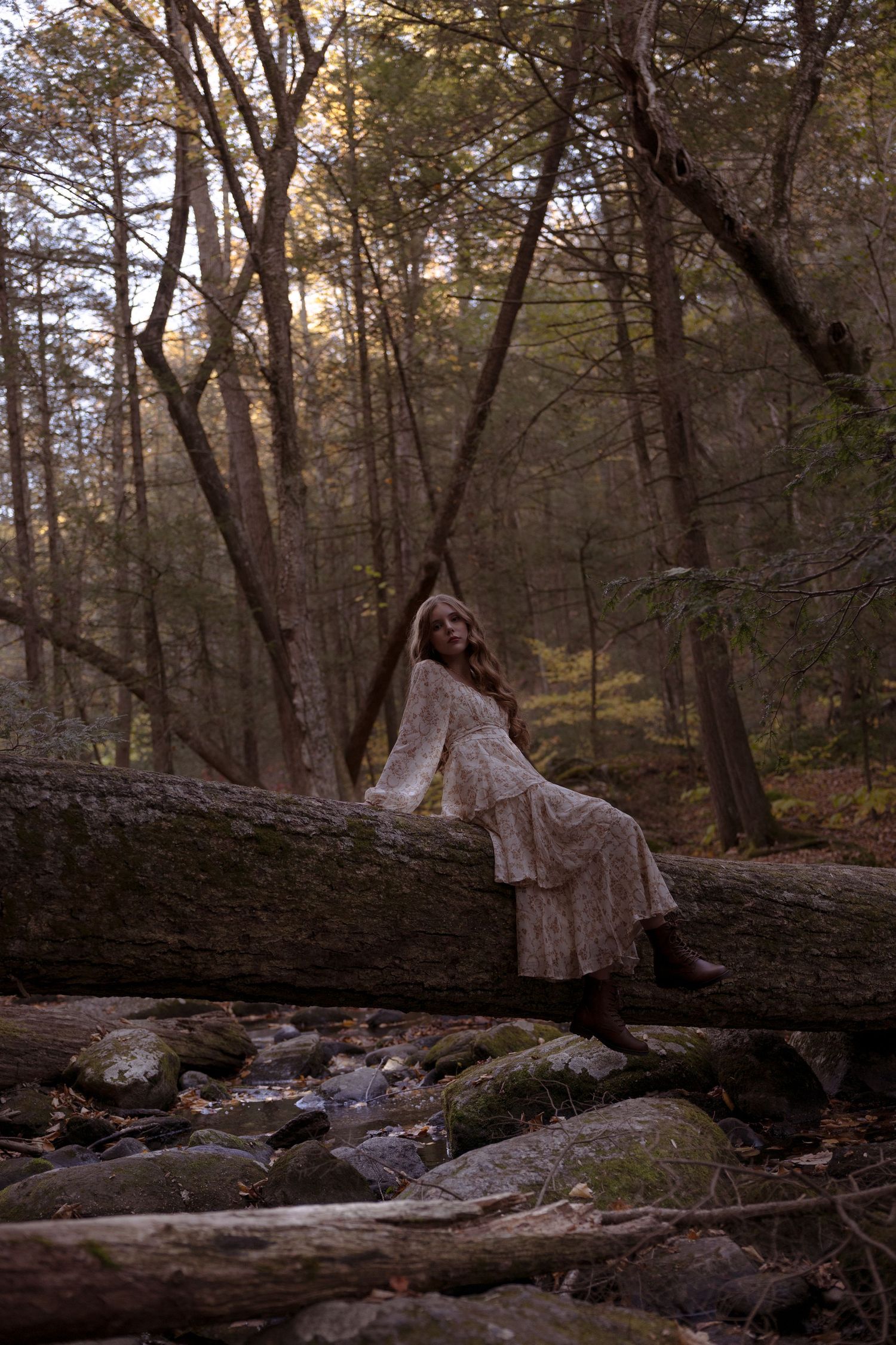 A person in a white dress sits on fallen logs in a moody autumn forest at sunset.