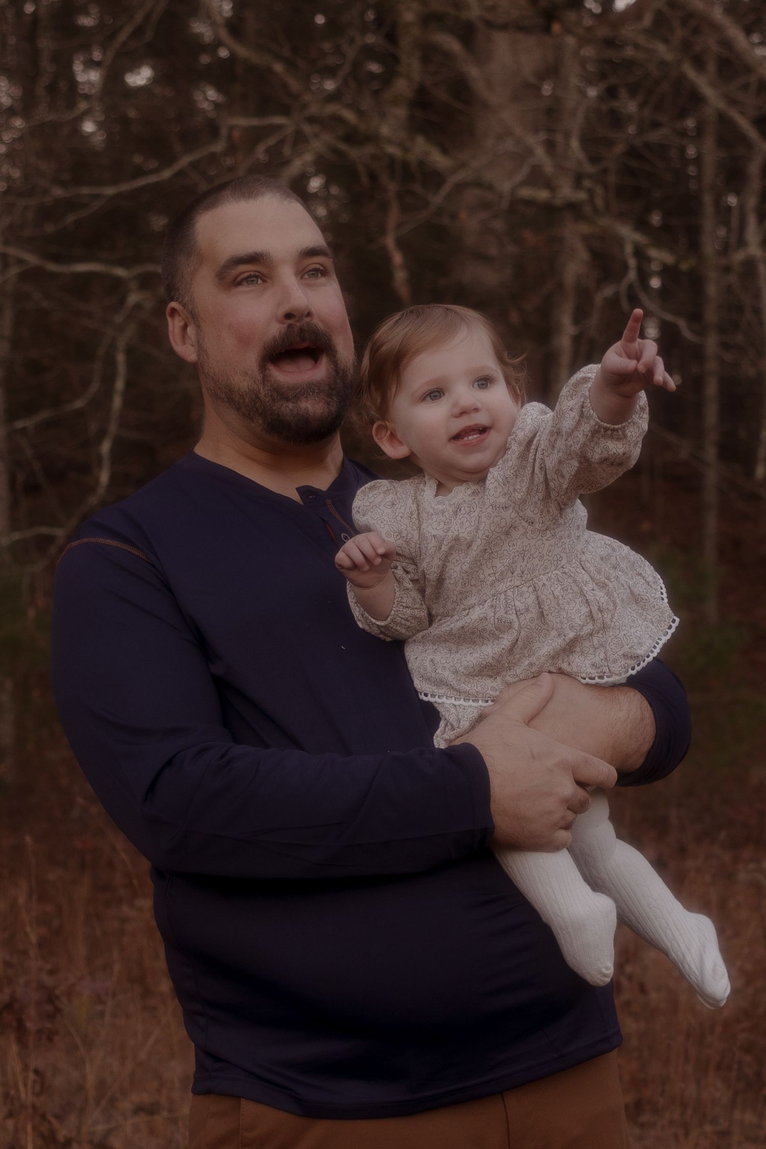 A person in a navy sweater holds and plays with a baby wearing a white lacy dress in a wooded outdoor setting.