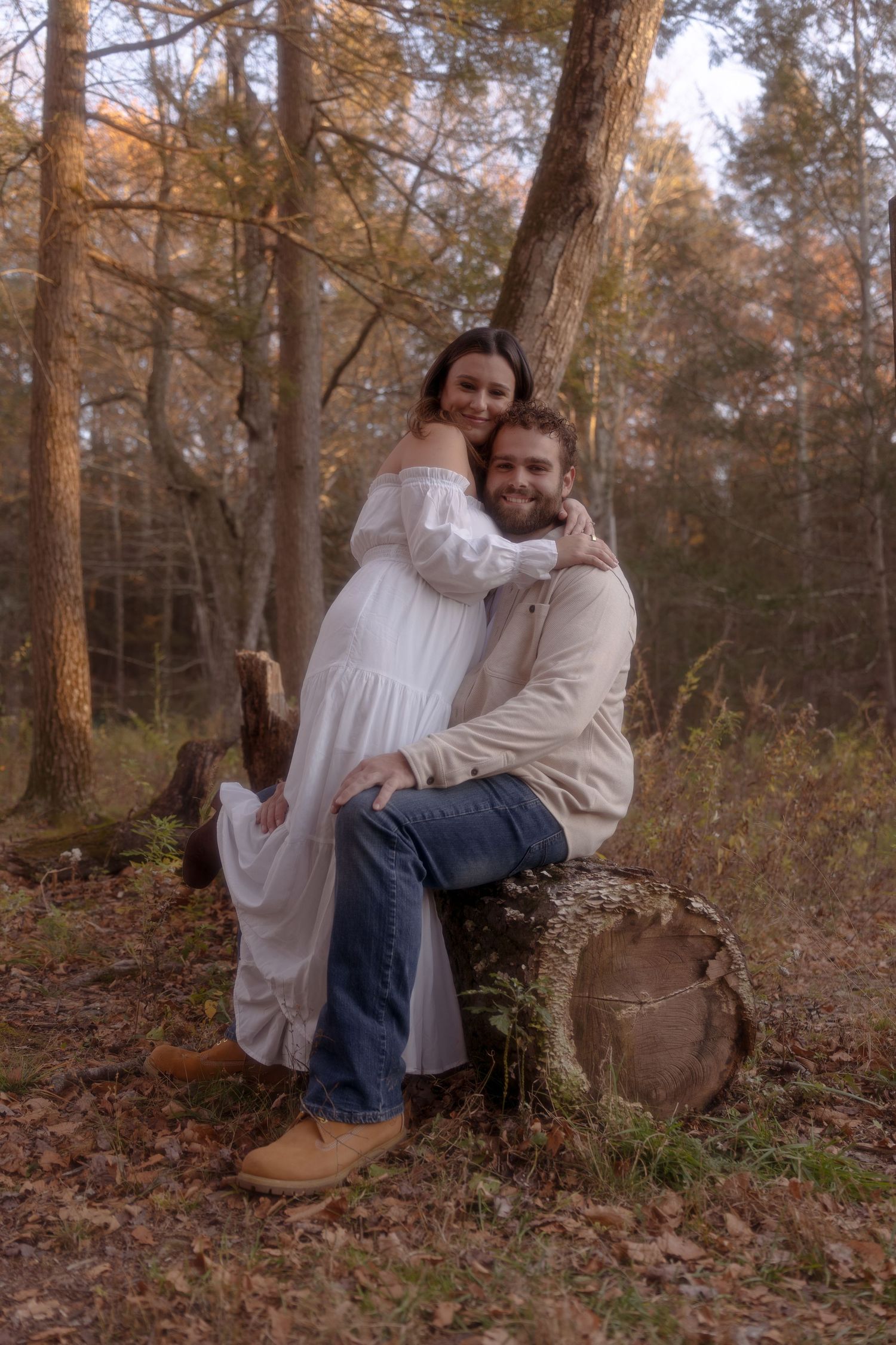 A couple sits together in a serene autumn forest setting with golden sunlight filtering through pine trees.